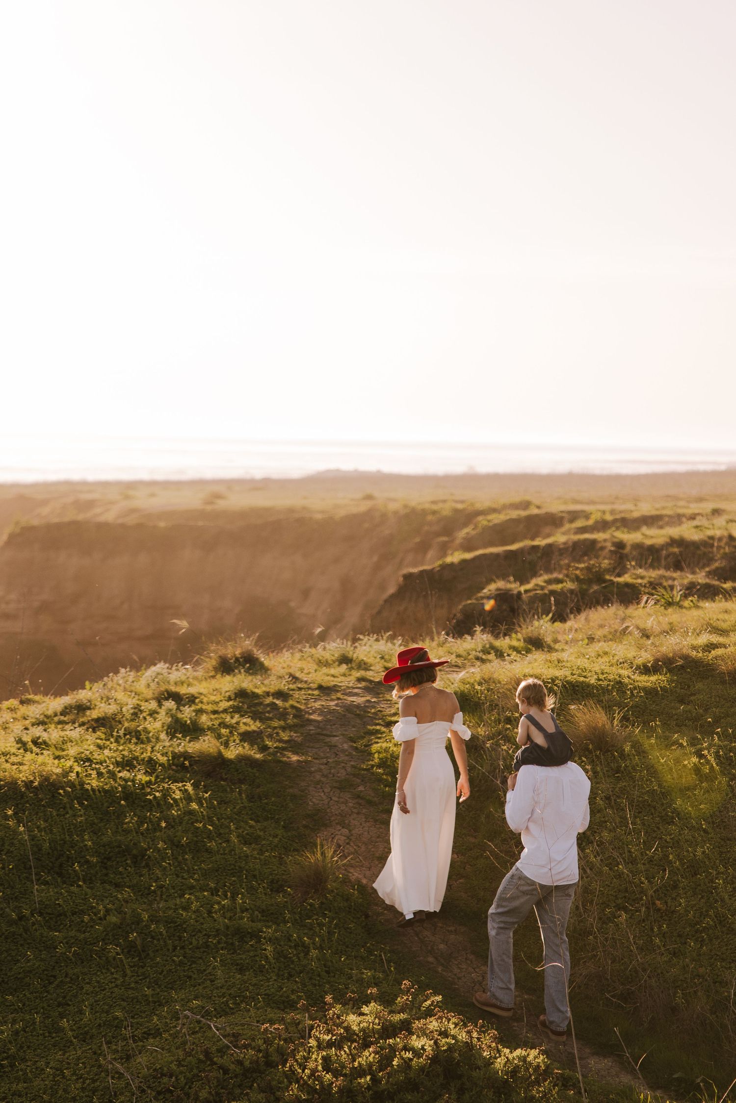 Photographer capturing a woman in a white dress and red hat on a scenic coastal cliff at golden hour.
