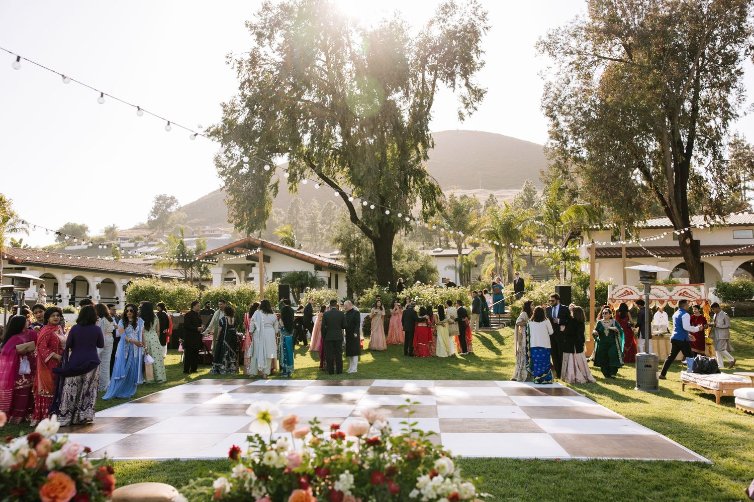 Outdoor Indian wedding ceremony on checkered dance floor surrounded by lush gardens.