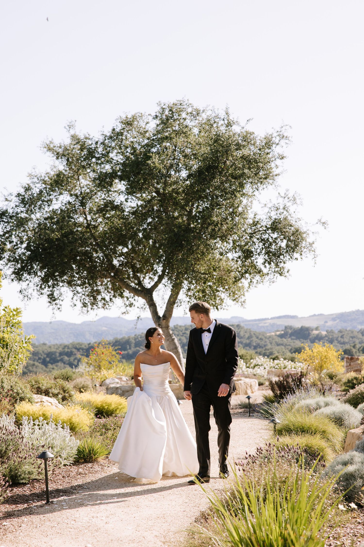 Bride and groom walk hand in hand along a garden path with sweeping mountain views behind them.