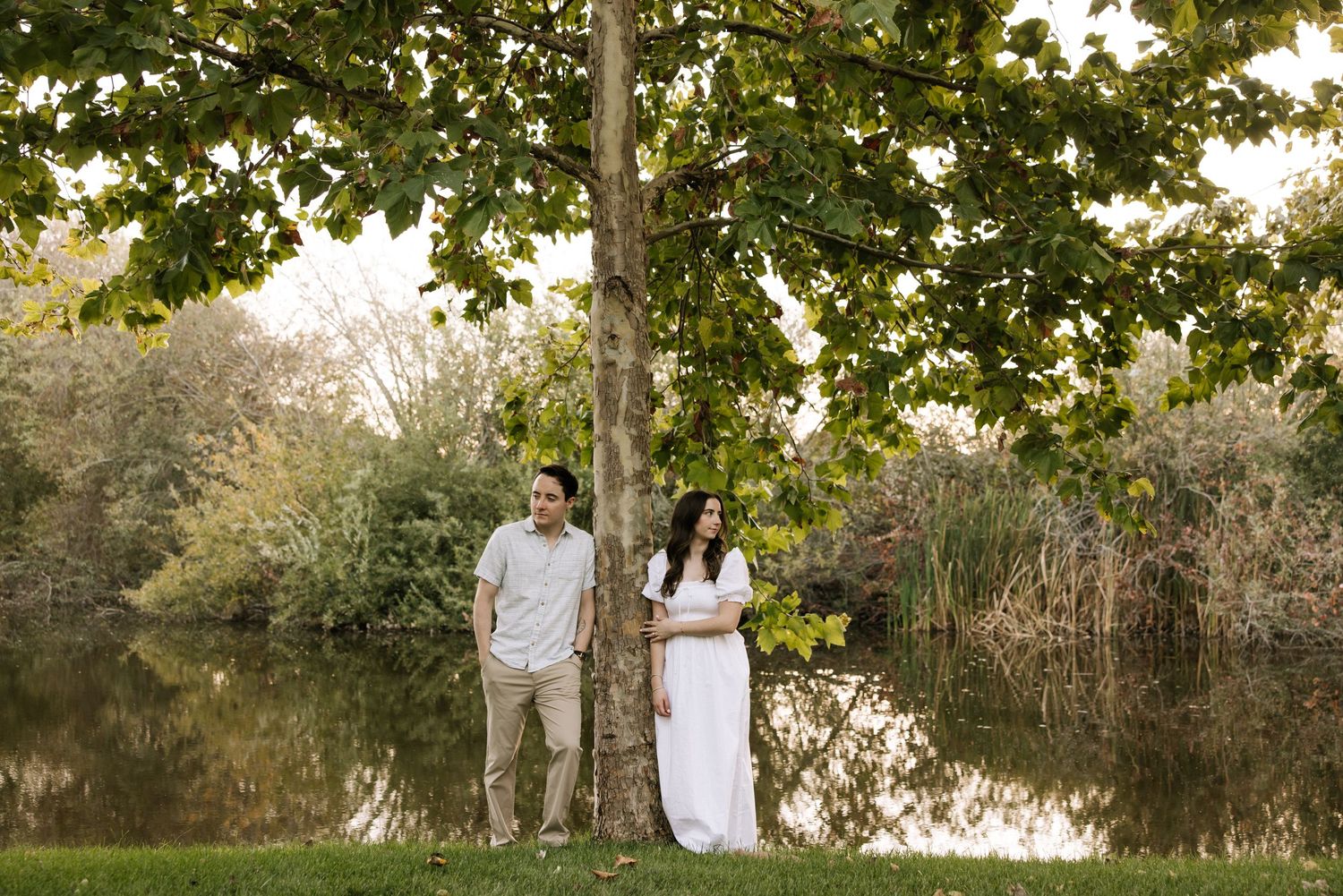 Couple standing by a tree near a serene pond in a lush green park, woman in white dress, man in casual attire.