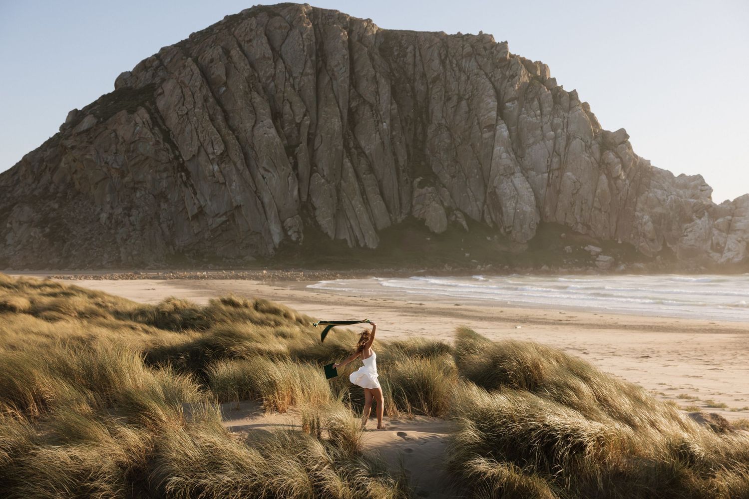 Woman dancing among coastal dunes with dramatic Morro Rock in background at golden hour.