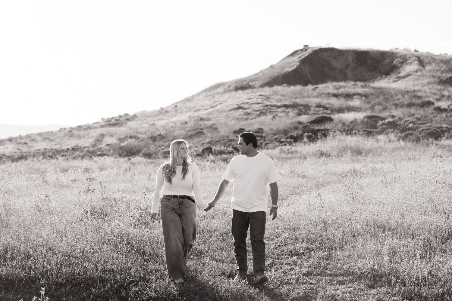Black and white photo of a couple walking hand in hand through a grassy hillside meadow.