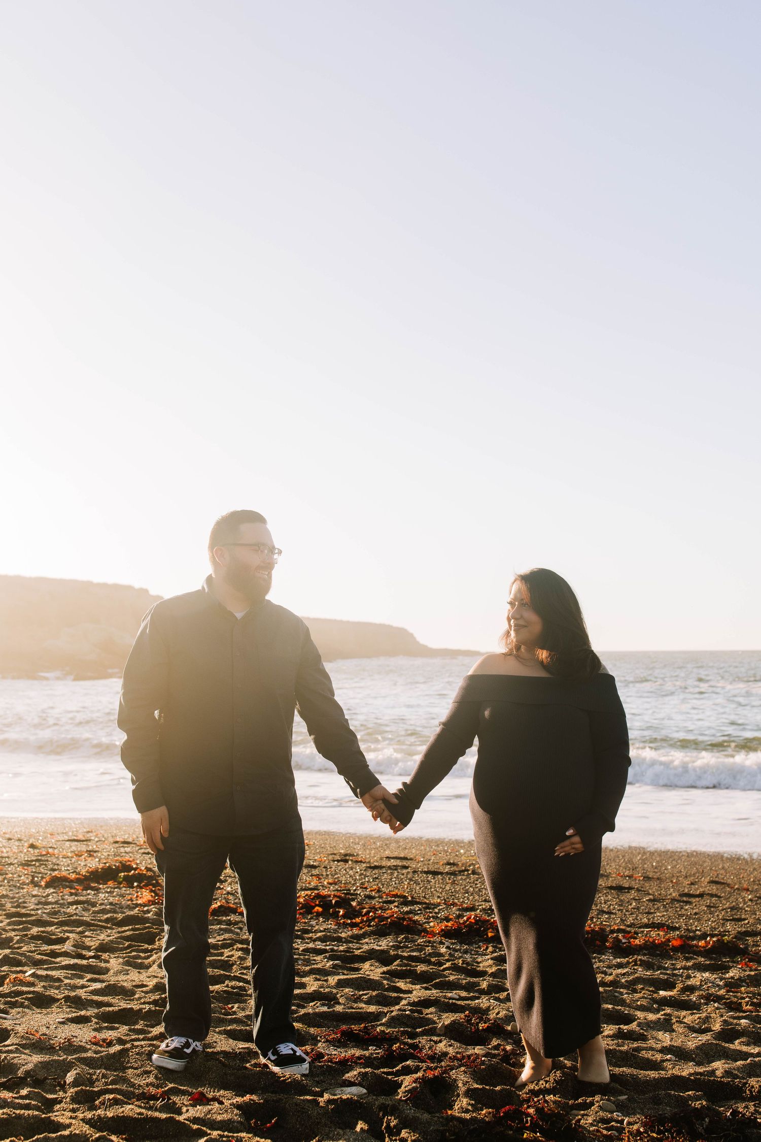 Couple holding hands on a sunlit beach, woman in black off-shoulder dress showing baby bump, golden hour light.