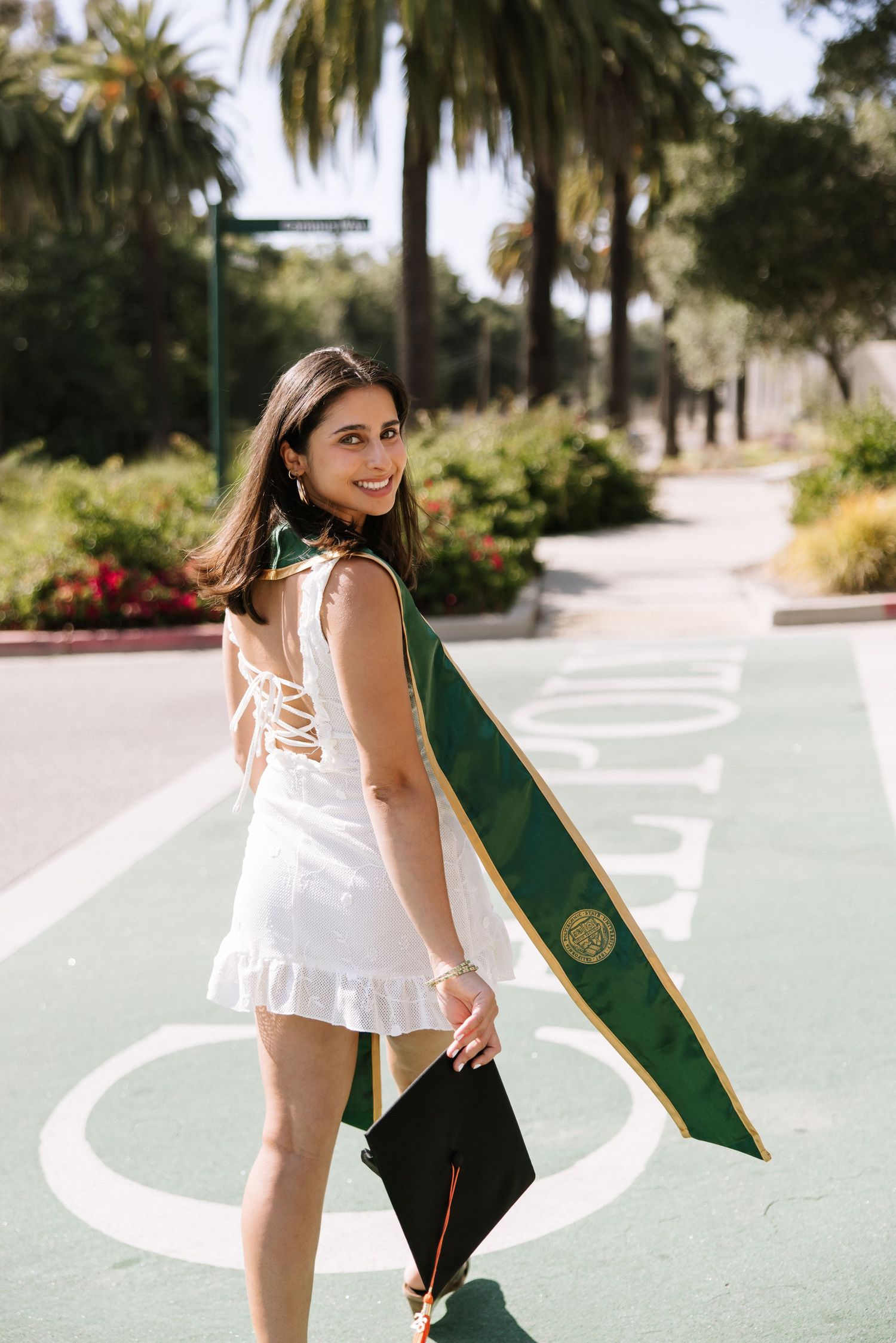 Graduate in white dress holding cap and green stole smiling on campus with palm trees.