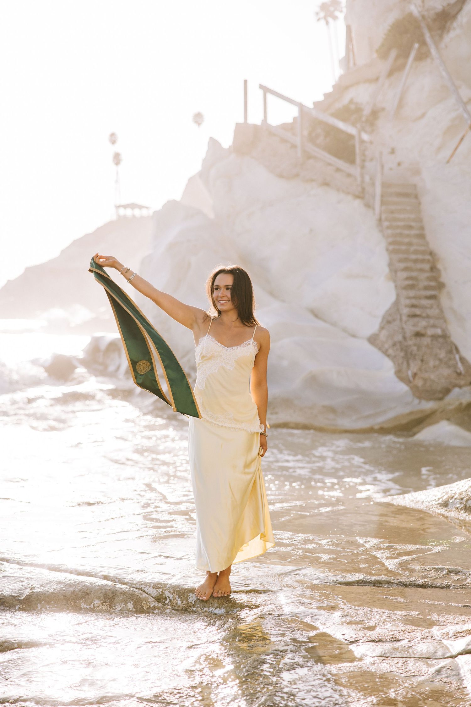 Woman in yellow dress holding green stole at rocky beach during golden hour.