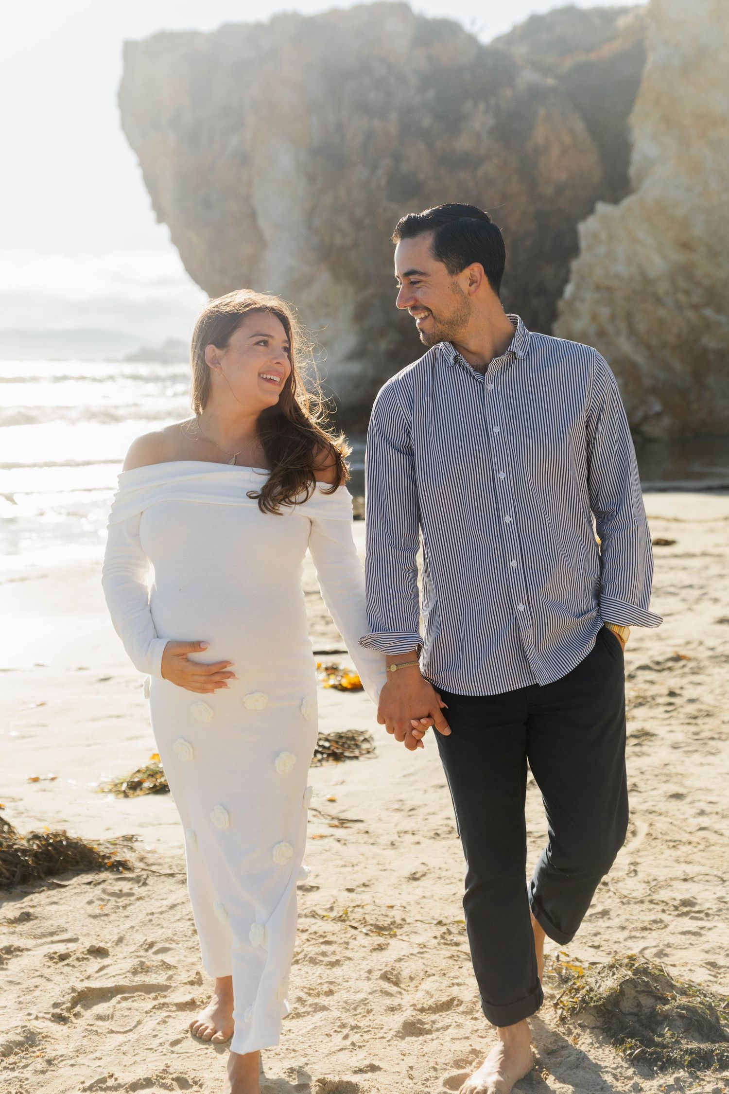 Happy pregnant couple walking hand in hand on sandy beach, woman in white off-shoulder maternity dress.