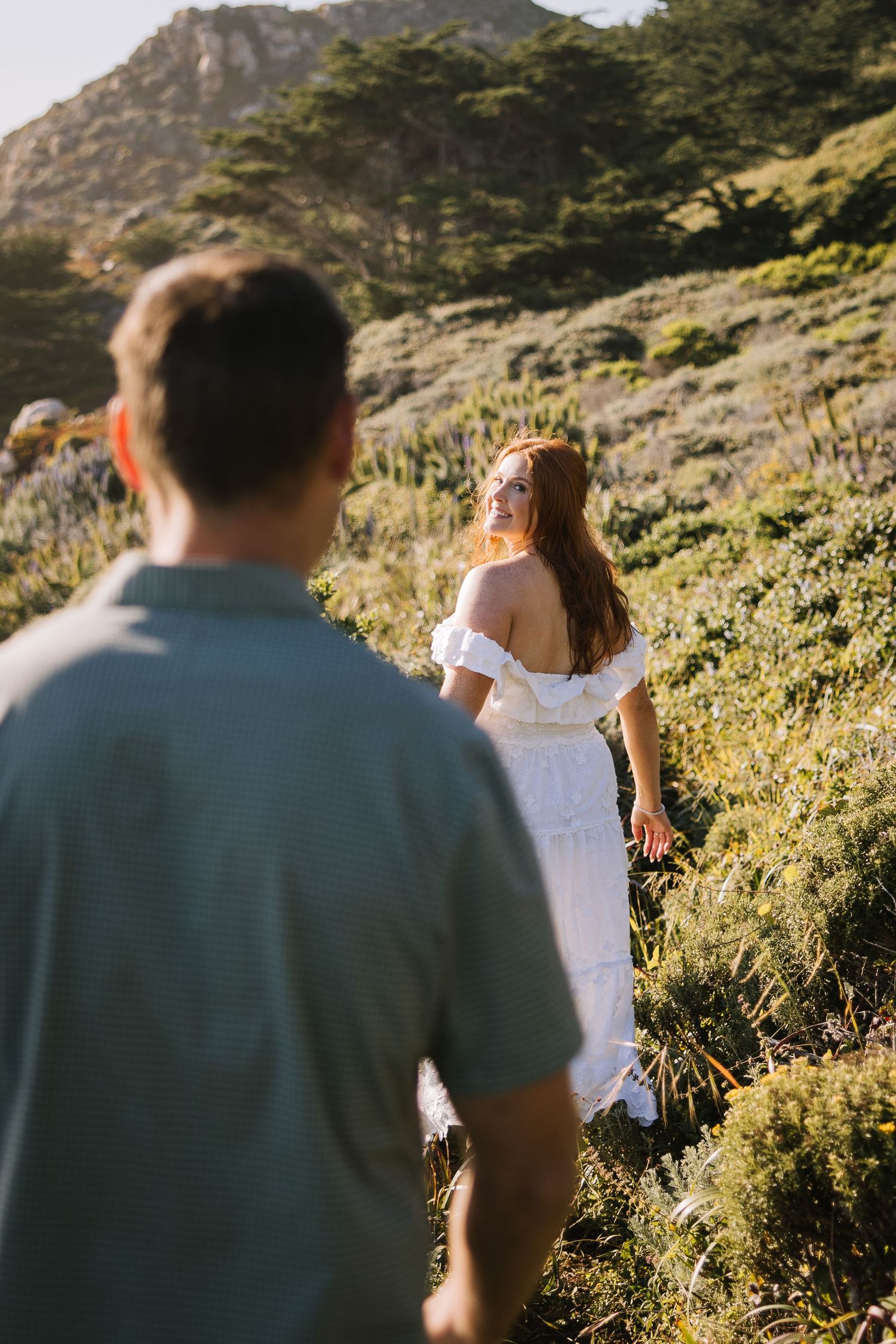 Woman in white off-shoulder dress looks back smiling at man following her up a sunlit hillside.