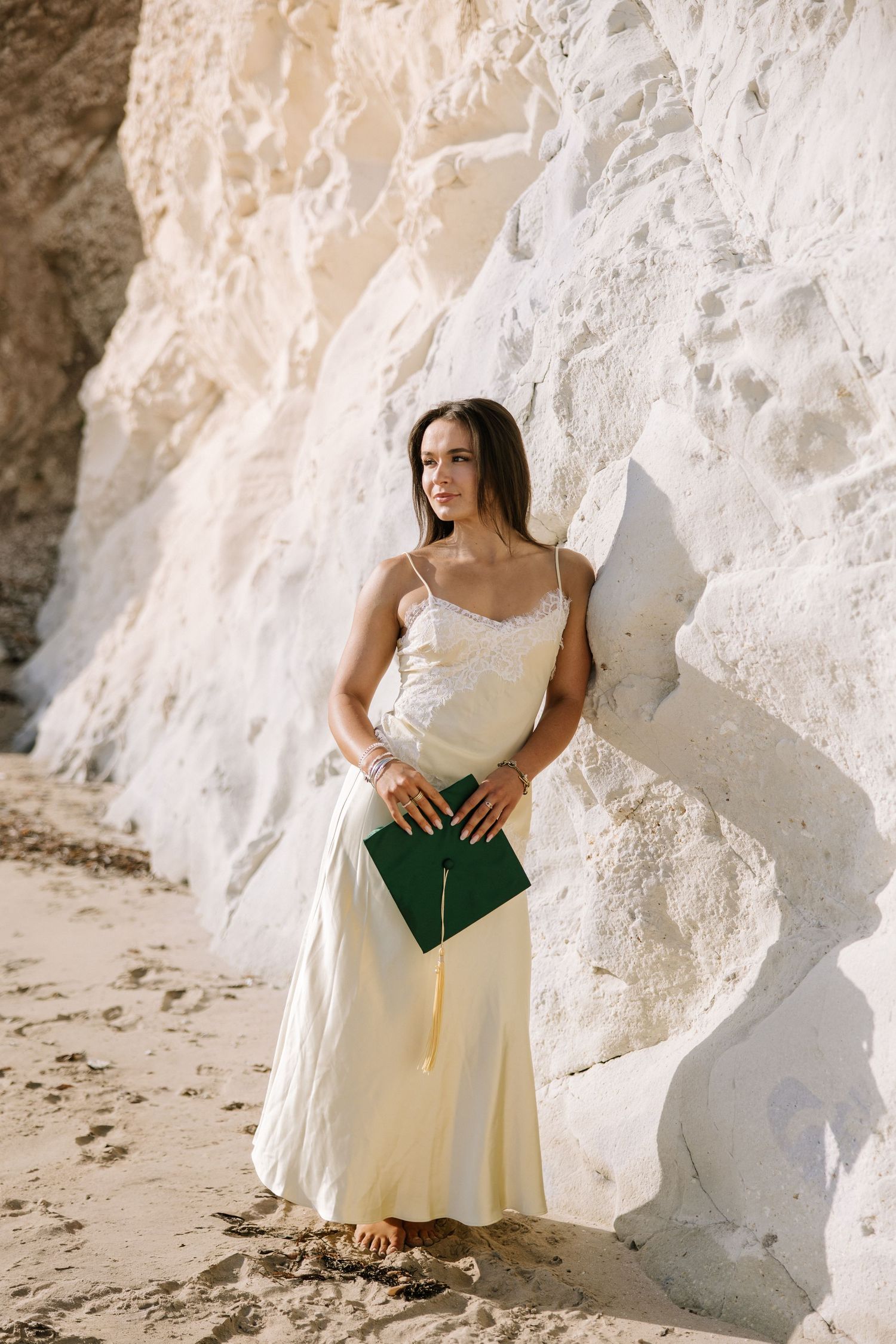 Elegant woman in cream slip dress holding a green graduation cap against a white sandy cliff.