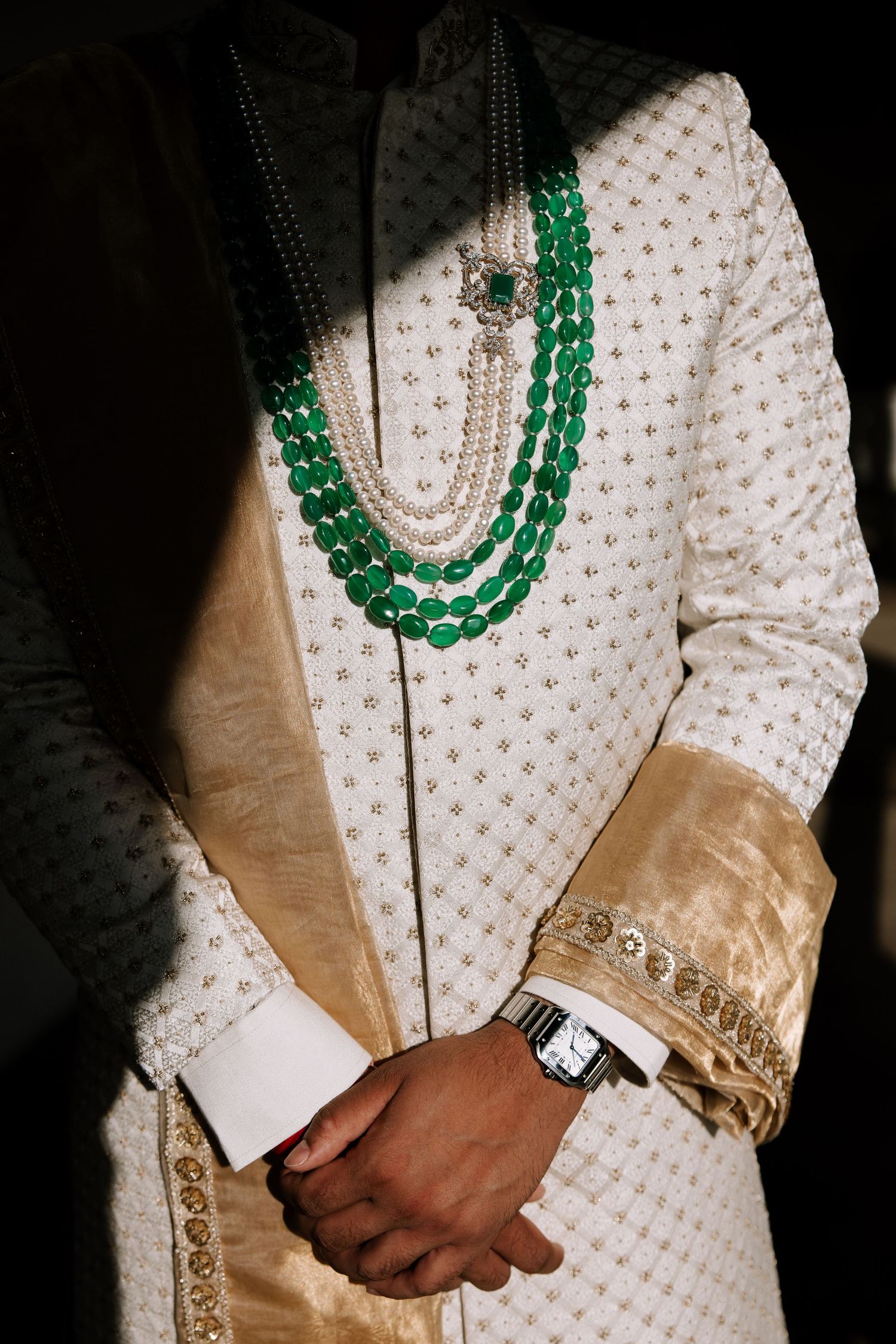 Groom in white sherwani adorned with emerald green and pearl layered necklaces at Indian wedding.