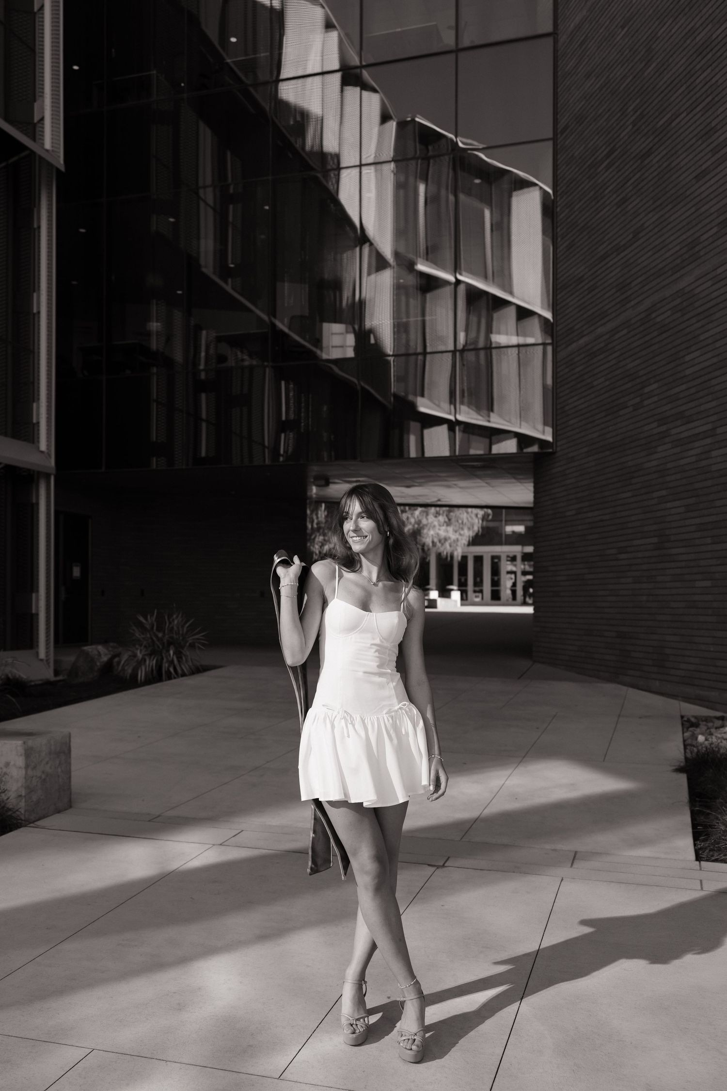 Young woman in white mini dress walks confidently past modern glass building in black and white portrait.