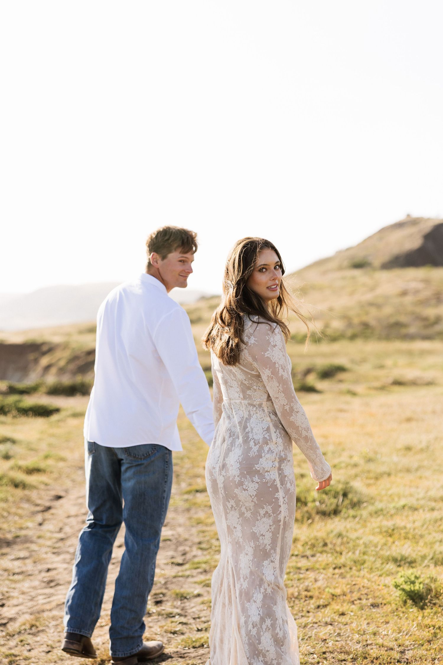 Couple holding hands walking through golden field at sunset, woman in lace dress looking back.