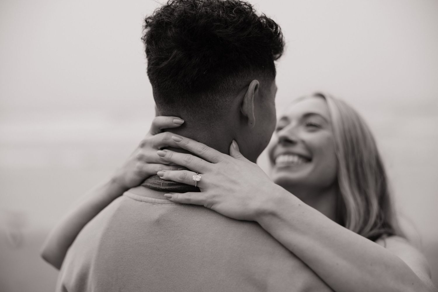 Close-up black and white photo of a smiling woman hugging a man, showing off her engagement ring.