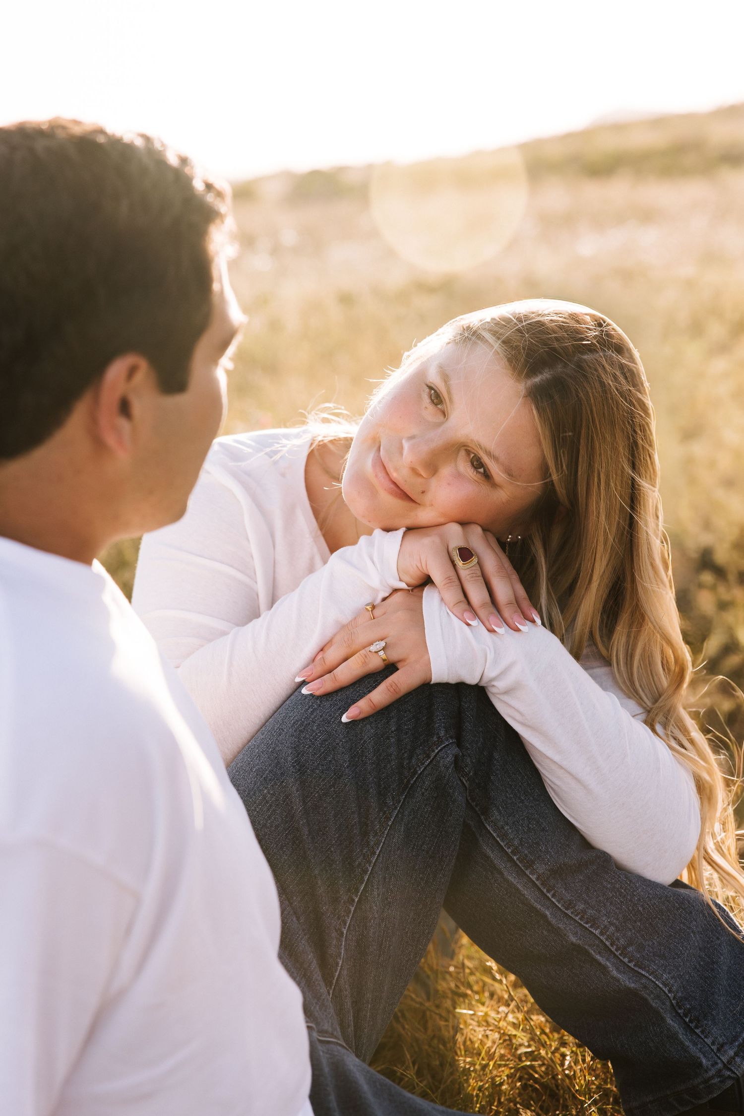 Woman gazes lovingly at partner while sitting in sunlit golden field during engagement shoot.