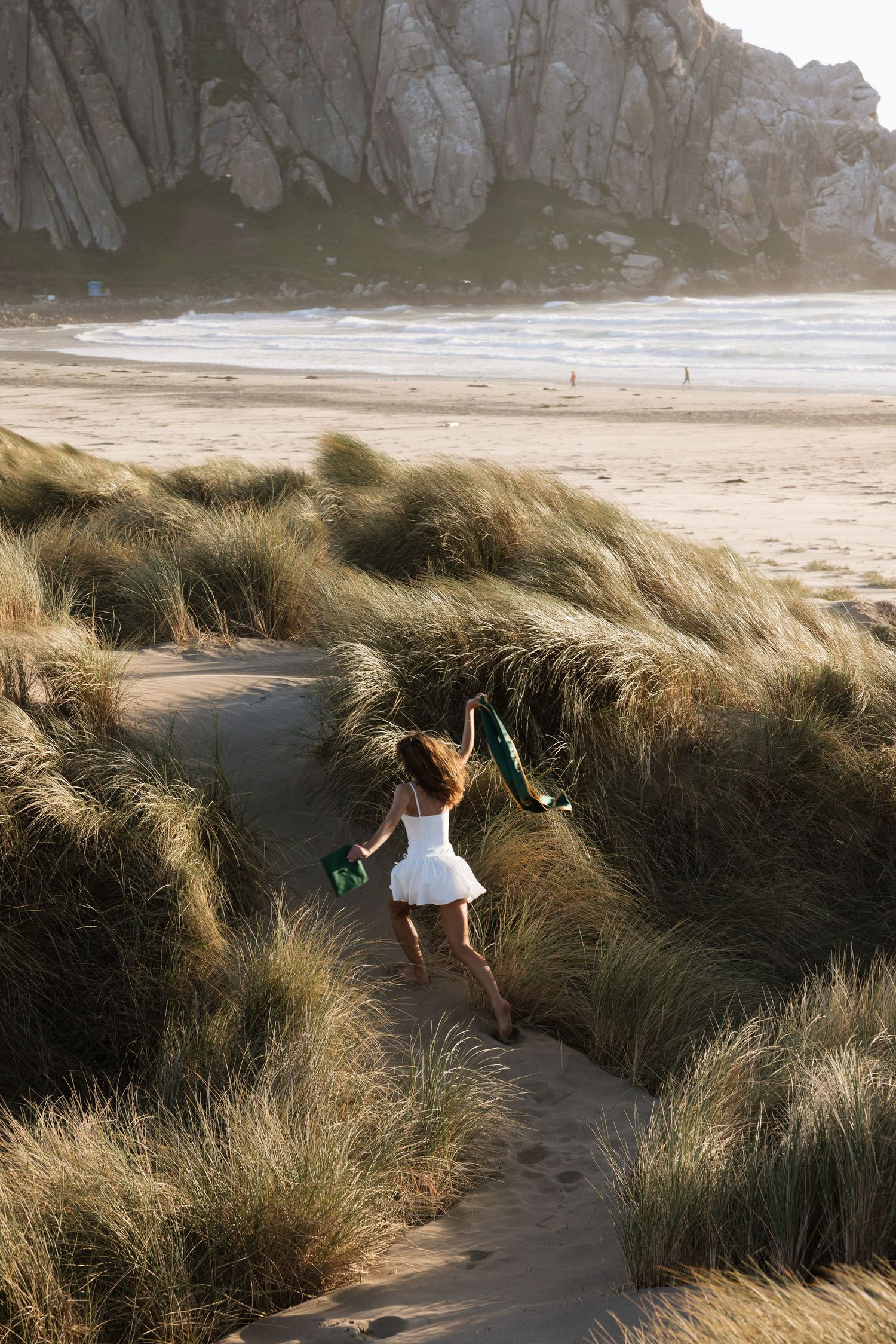 Woman in white dress walking through coastal sand dunes carrying green graduation sash toward ocean beach.