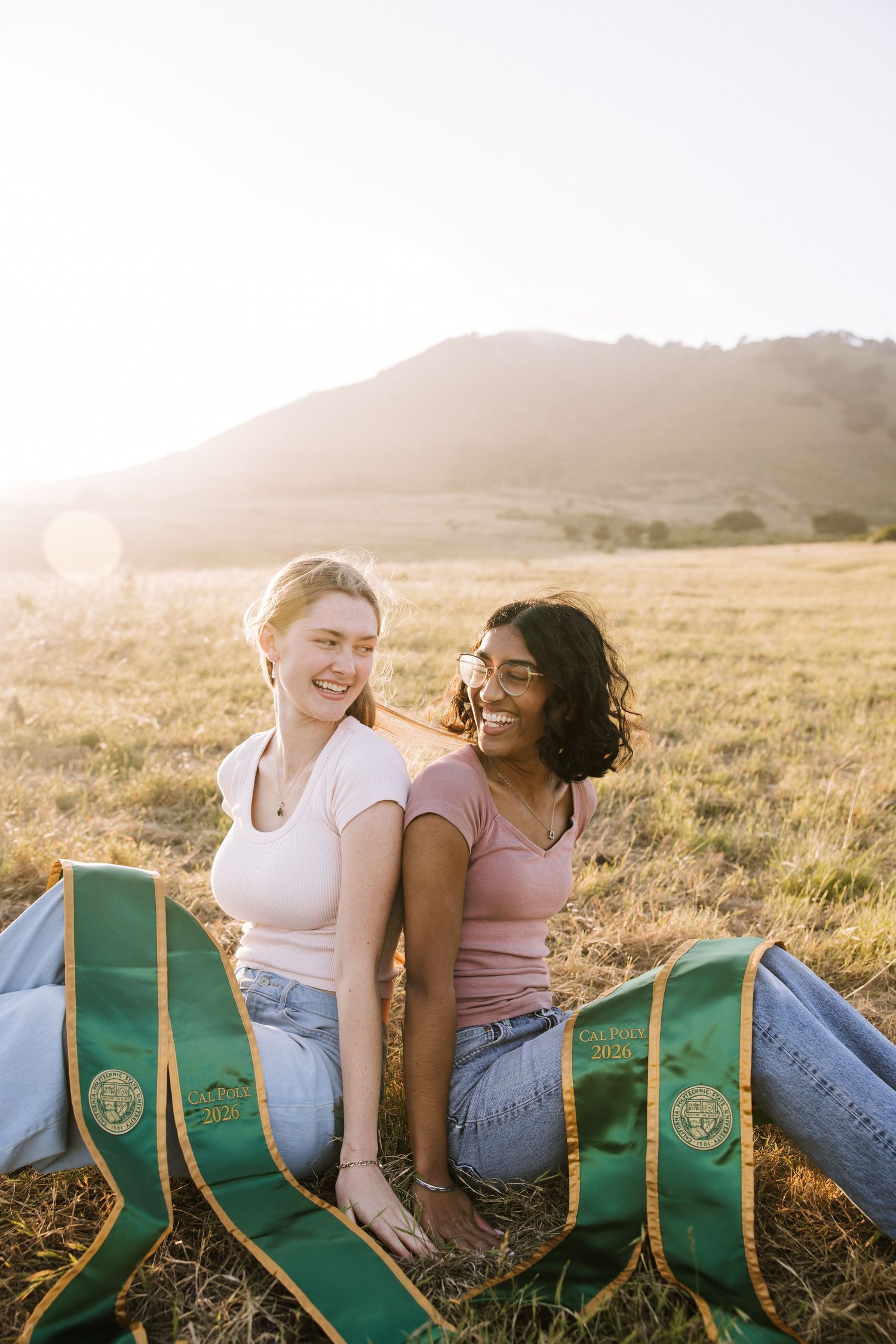 Two college graduates sit back-to-back in golden field at sunset with green graduation stoles.