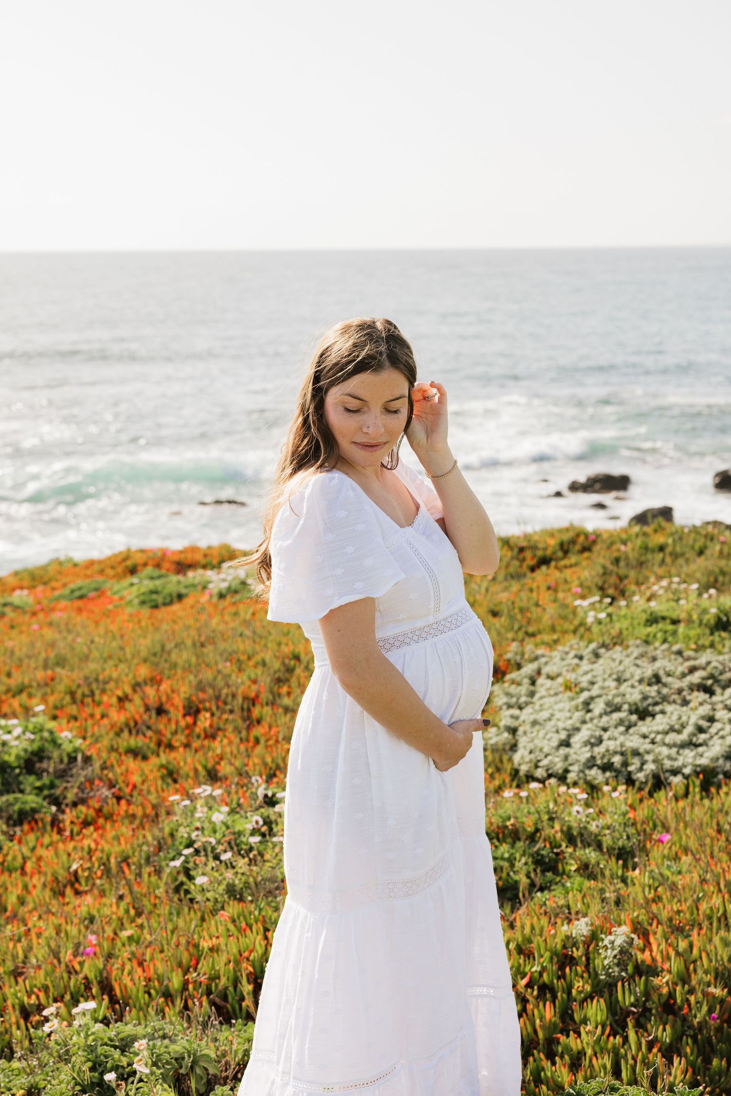Pregnant woman in white dress holding belly near colorful coastal wildflowers with ocean backdrop.