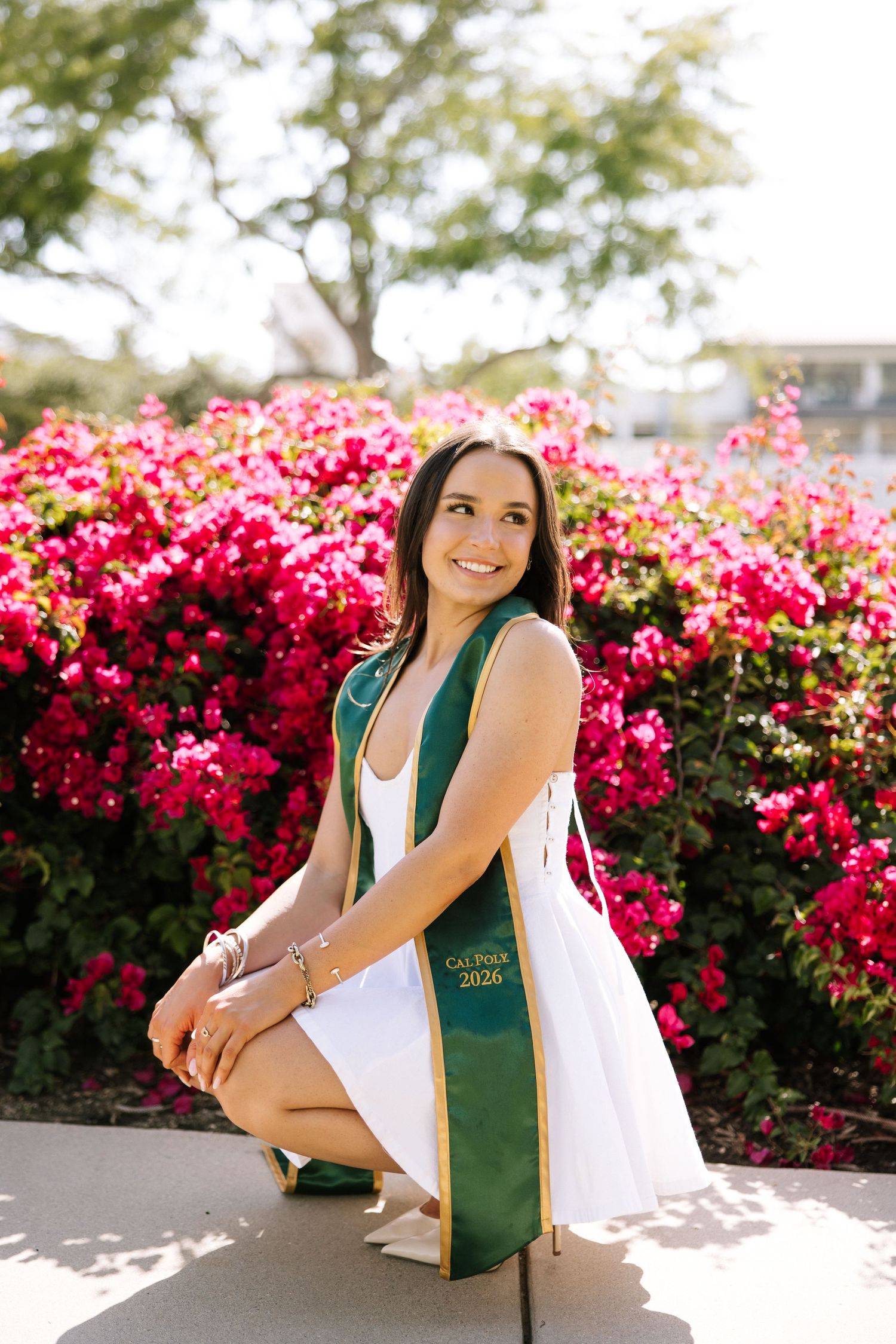 Smiling graduate in green Cal Poly 2026 stole posing before vibrant pink bougainvillea flowers.