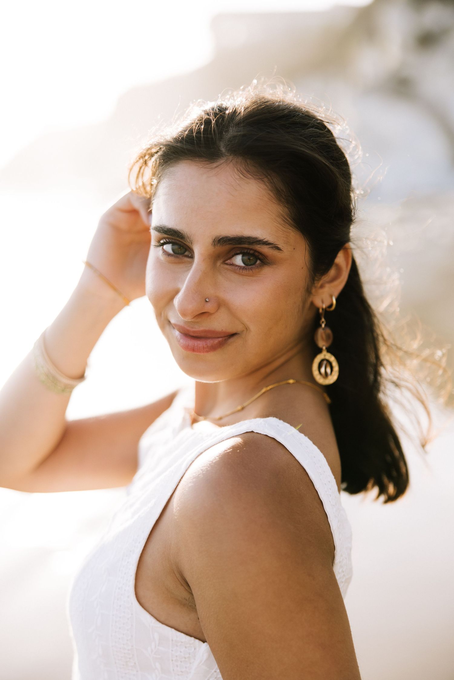 Close-up portrait of young woman with gold earrings smiling in warm sunlight outdoors.