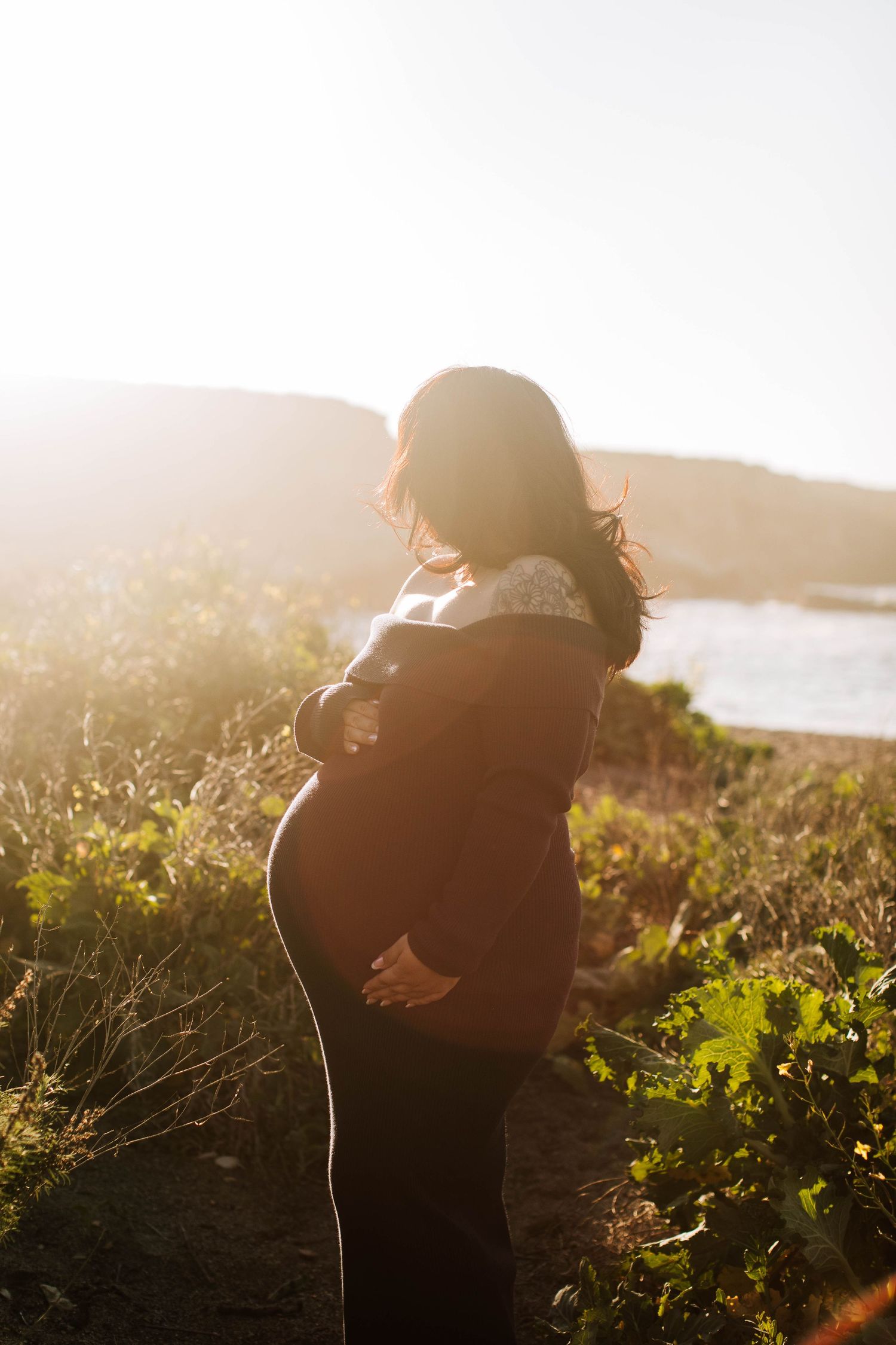 Backlit maternity photo of pregnant woman holding belly in golden sunset light outdoors.