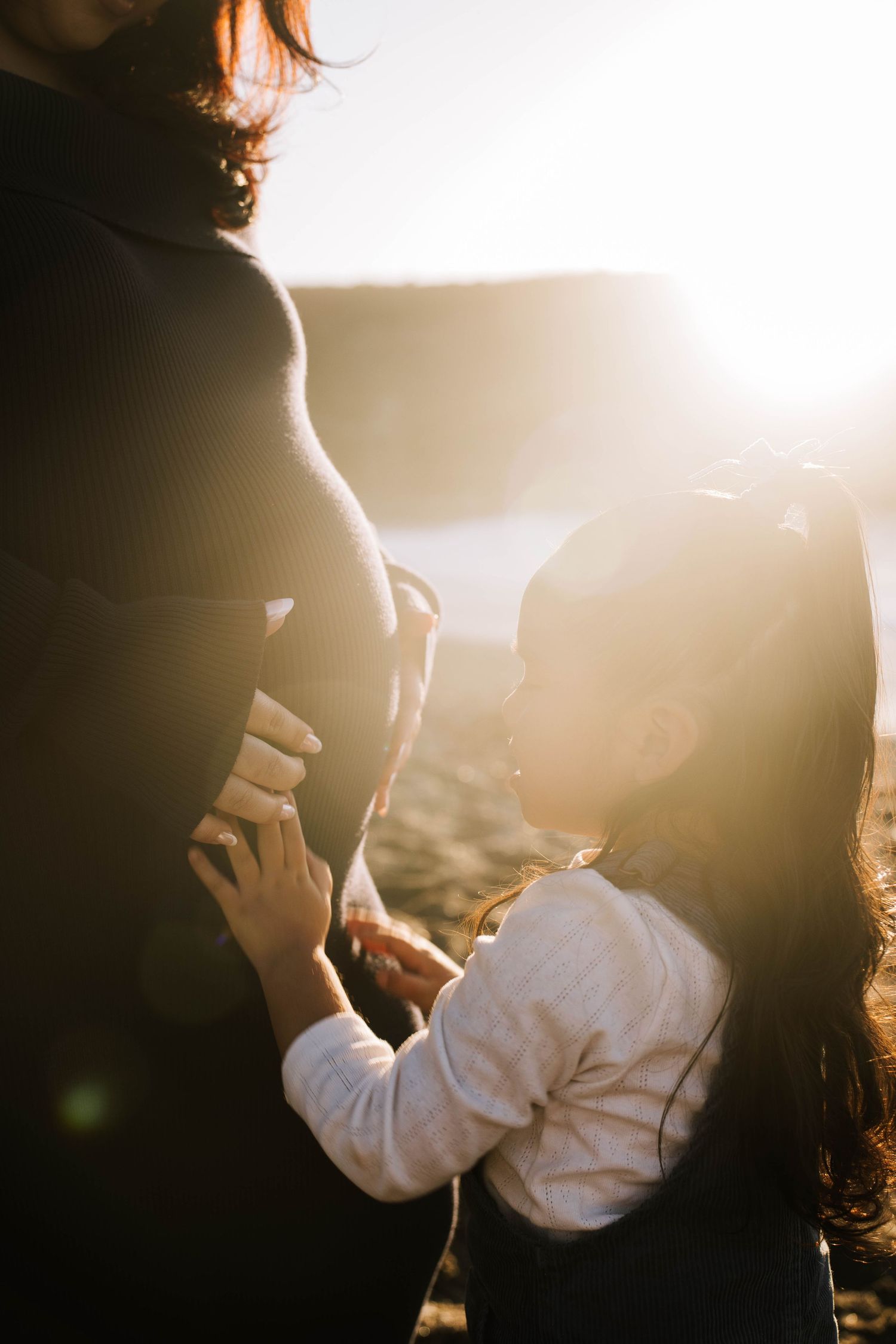 Young girl gently touching pregnant mothers belly in warm golden sunset light on a beach.