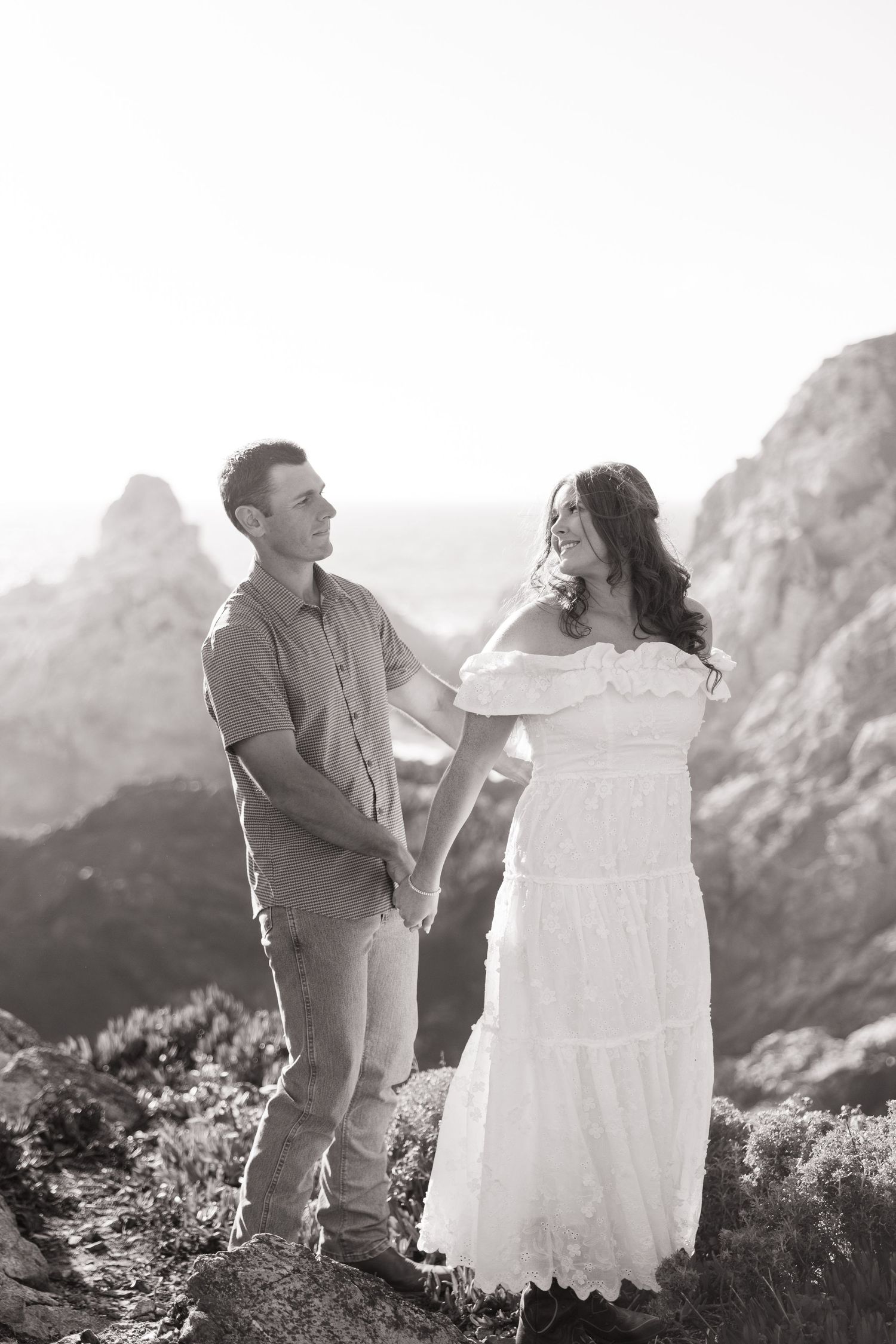Black and white portrait of couple holding hands and gazing at each other on rocky coastal cliffs.