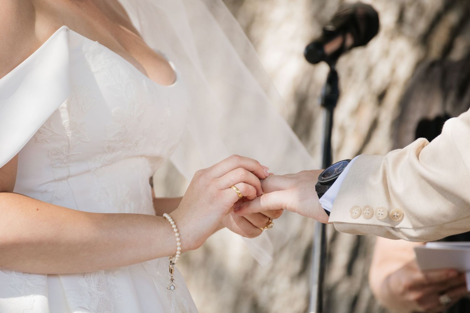 Bride placing a wedding ring on groom's finger during outdoor ceremony.