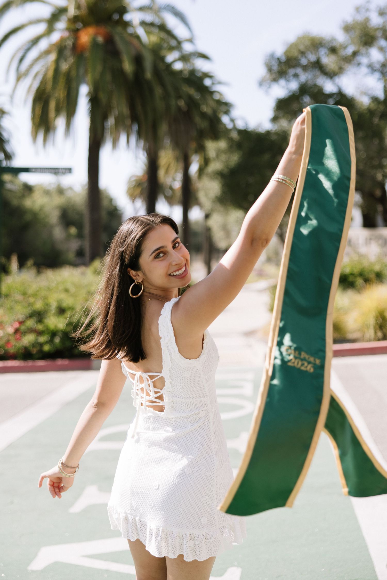 Joyful graduate in white dress raising green 2026 stole on sunny campus with palm trees.