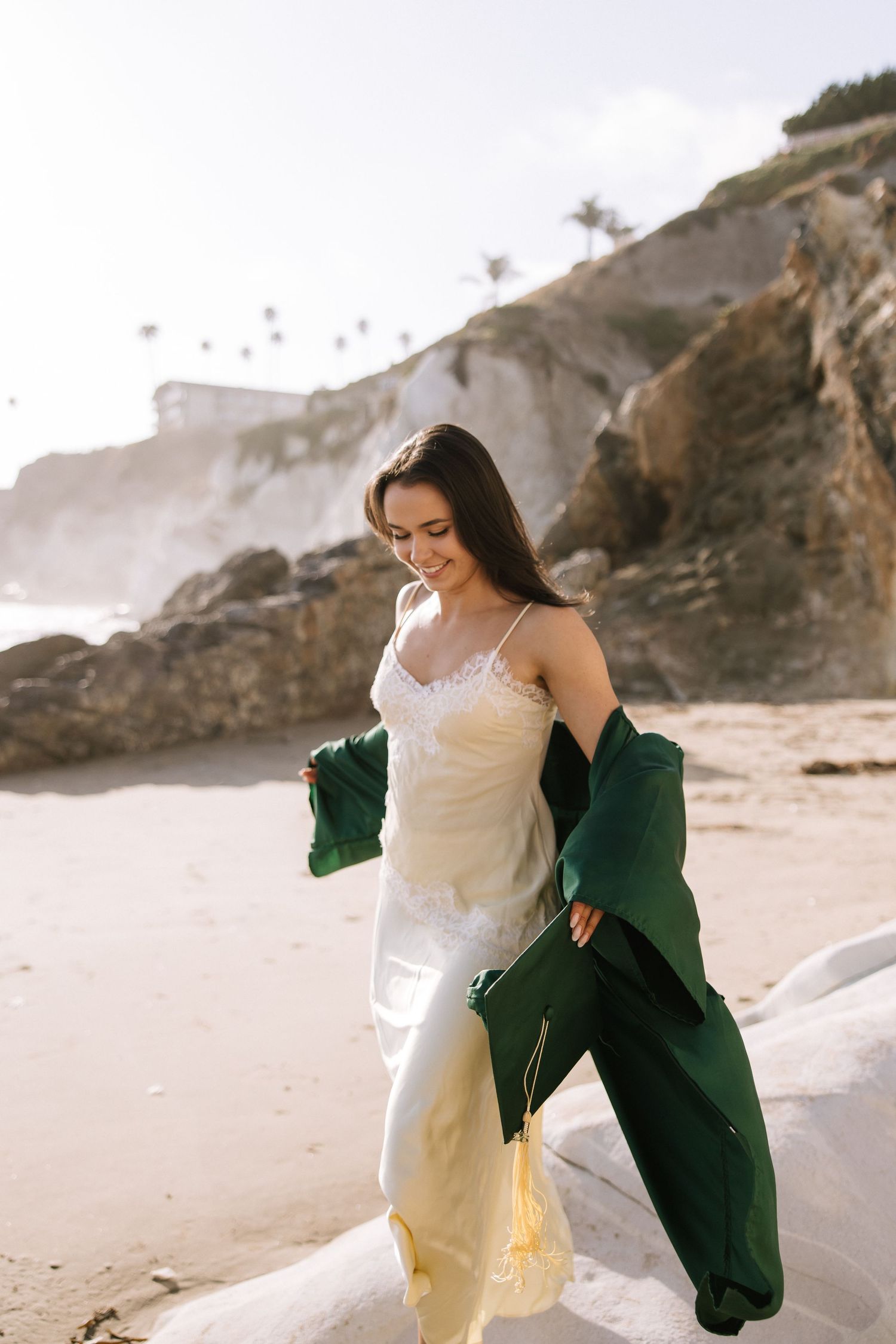 Woman in yellow slip dress holding green graduation stole on sunlit rocky California beach.
