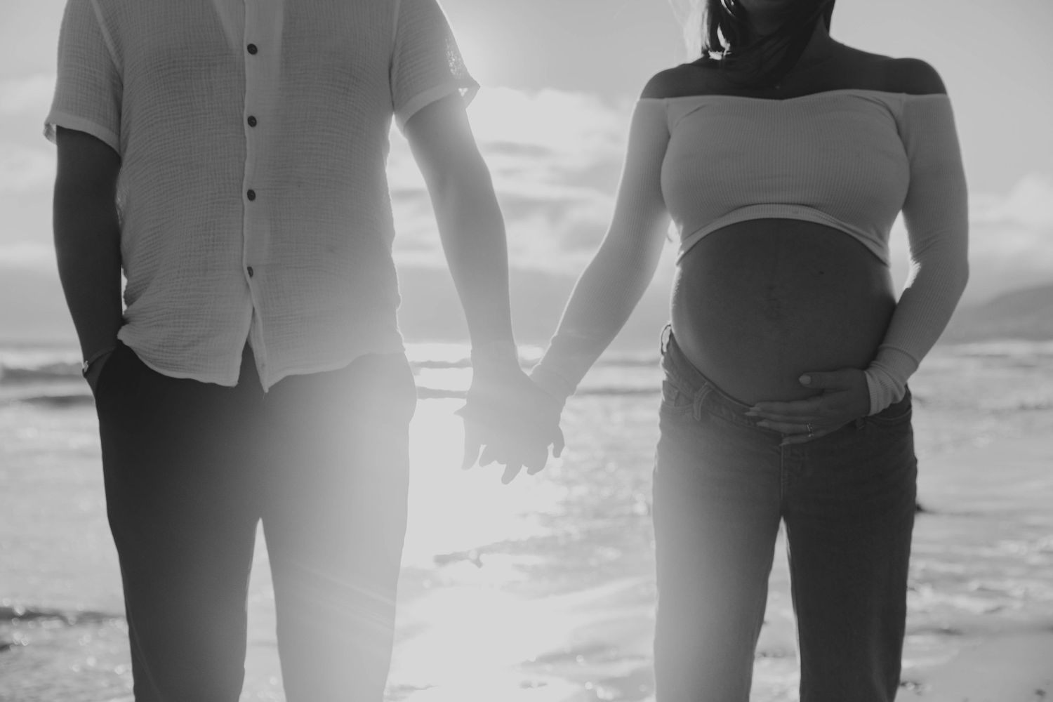 Black and white maternity photo of a couple holding hands on a sunny beach, woman cradling her baby bump.