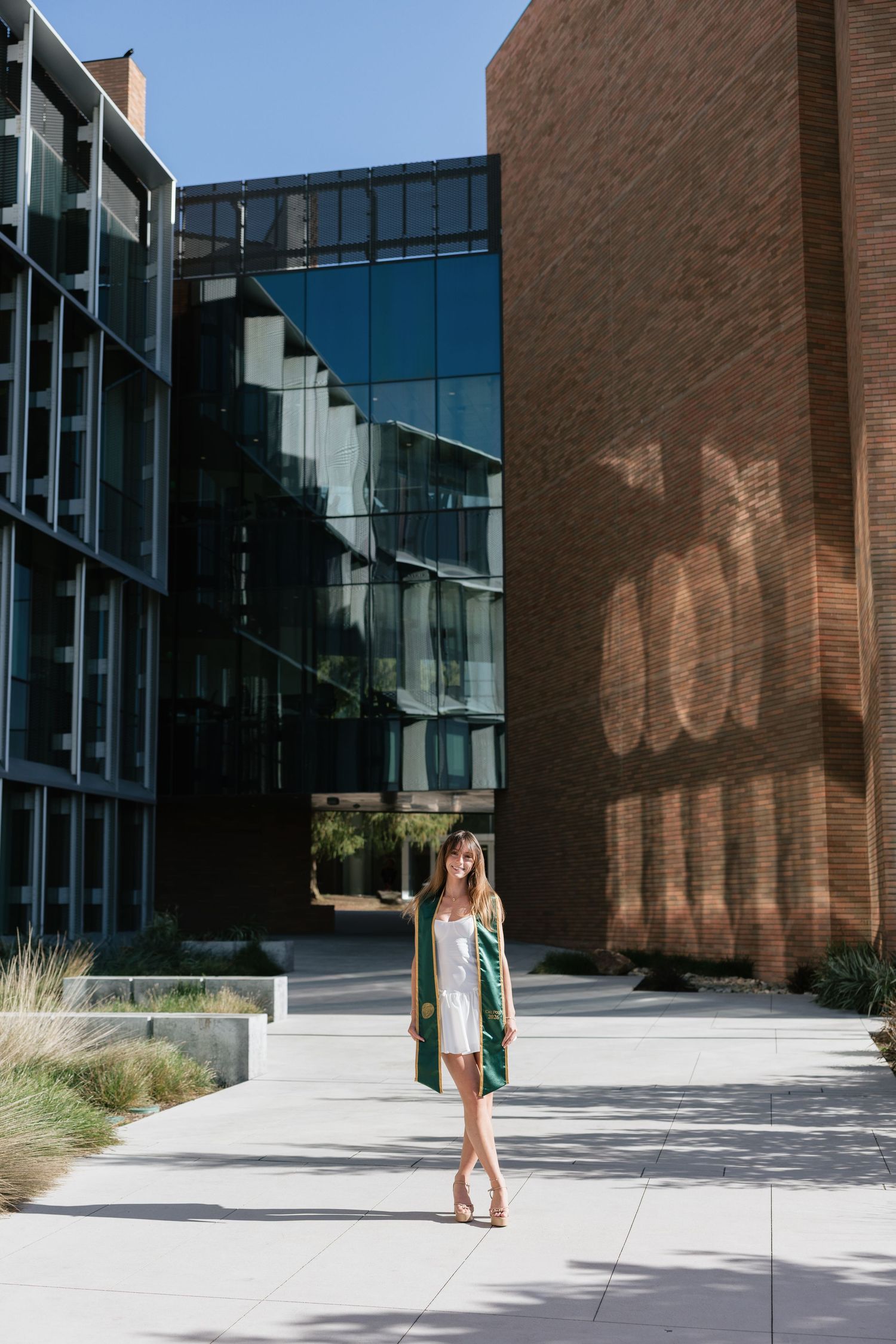 Young woman in green graduation gown and white dress standing outside modern university building.