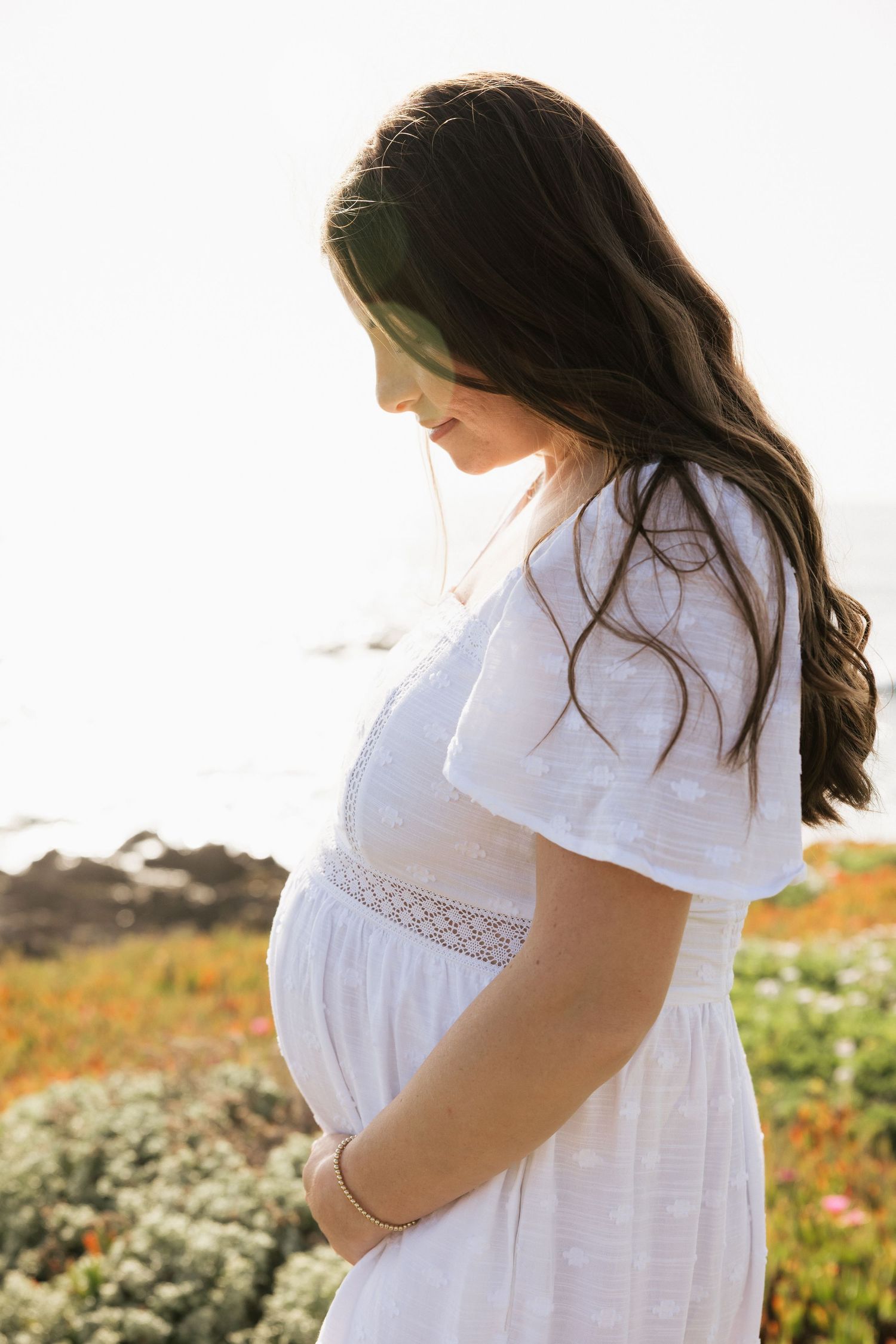 Side profile of pregnant brunette woman in white dress cradling belly near ocean wildflowers.