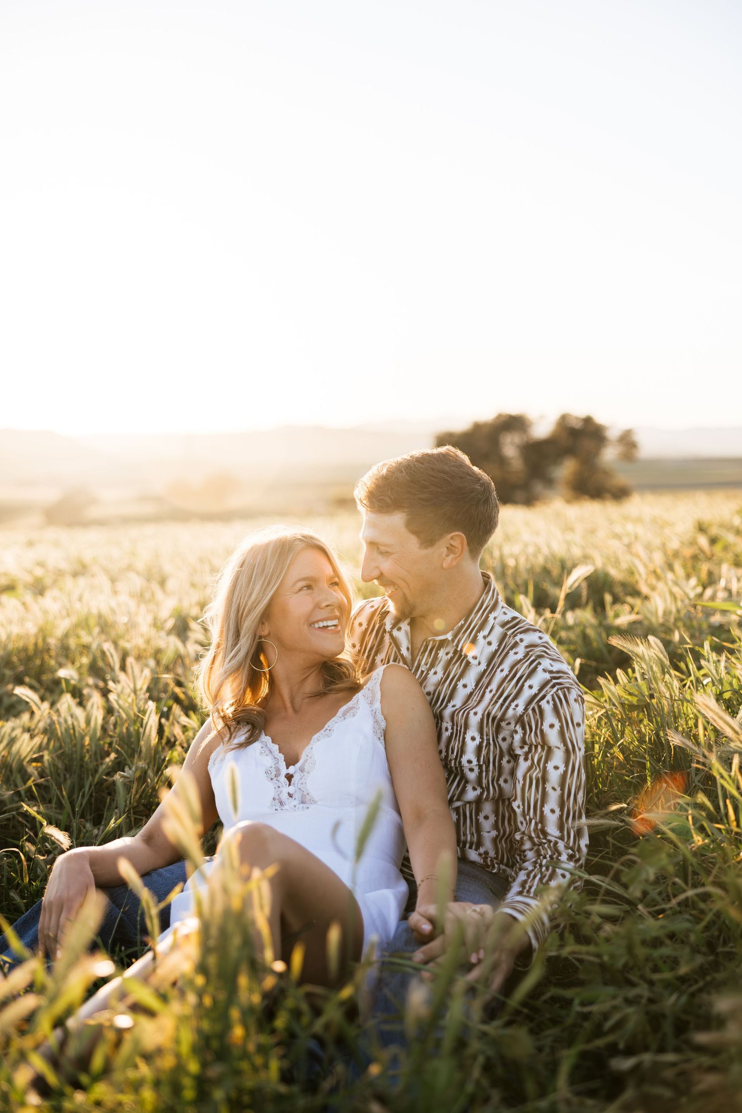 Couple sitting together in tall grass field smiling at each other during sunset.