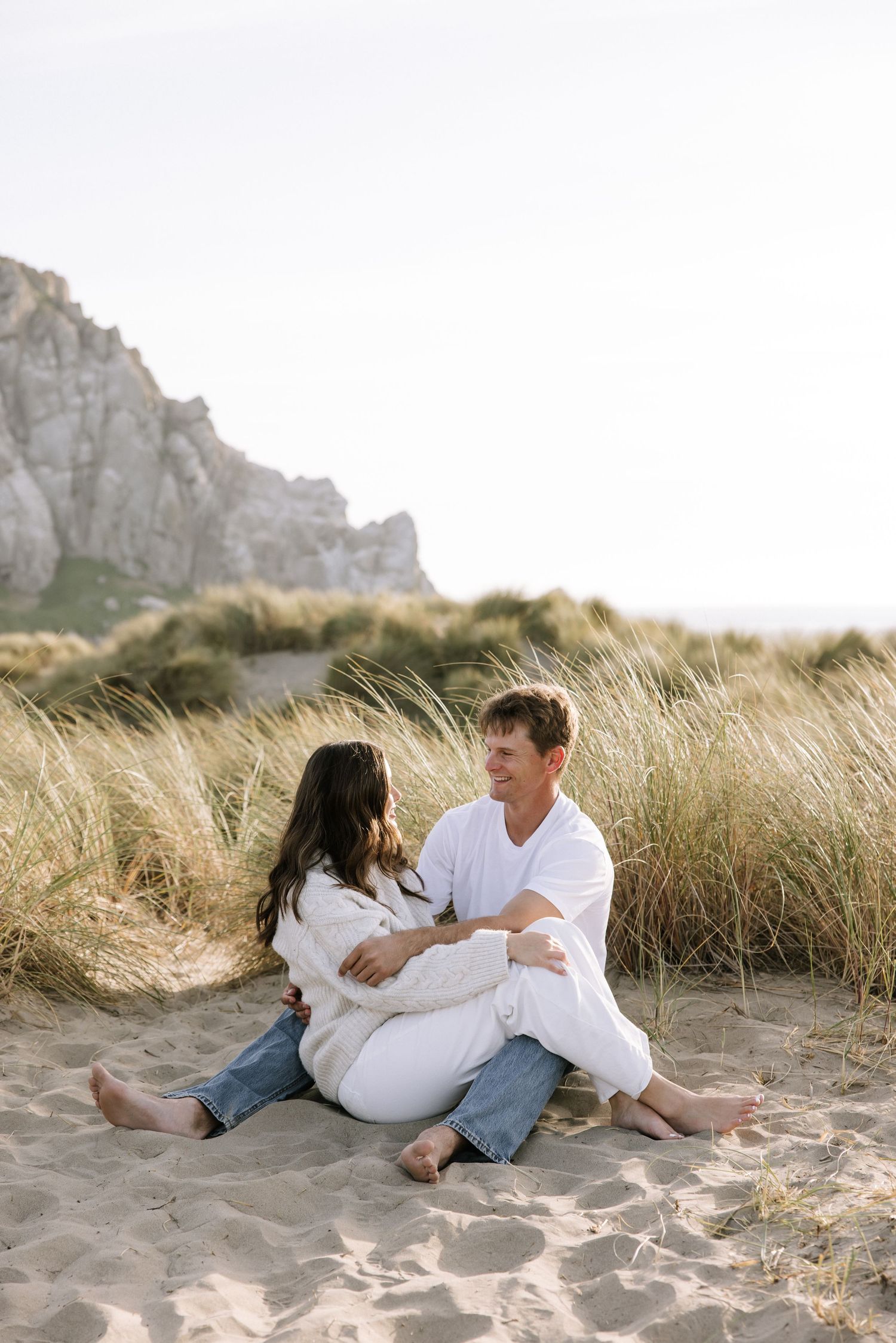 Couple sitting together in sandy beach dunes with dramatic rock formation backdrop on sunny day.