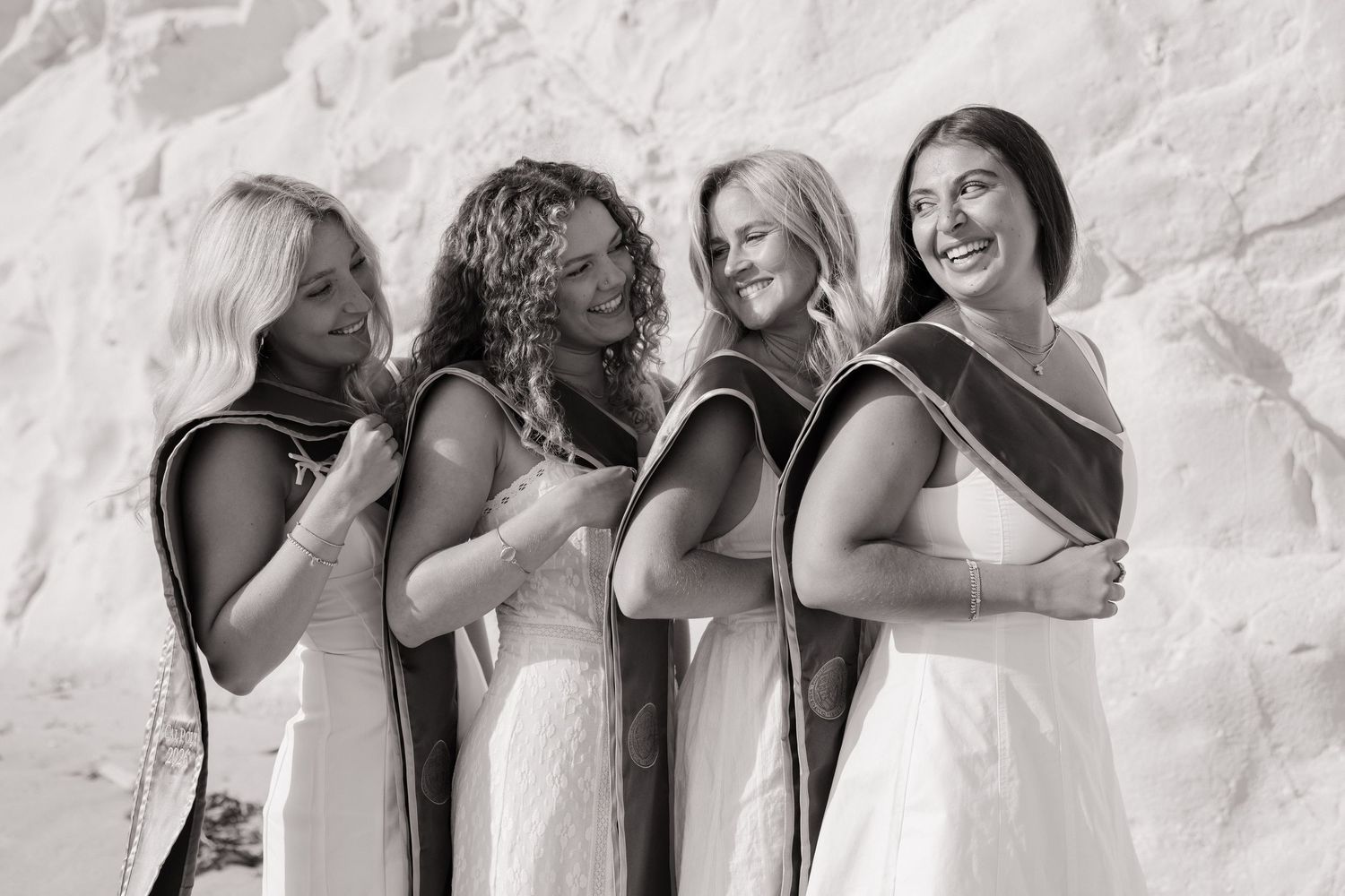 Four smiling female graduates in white dresses and sashes laugh together at beach in black and white photo.