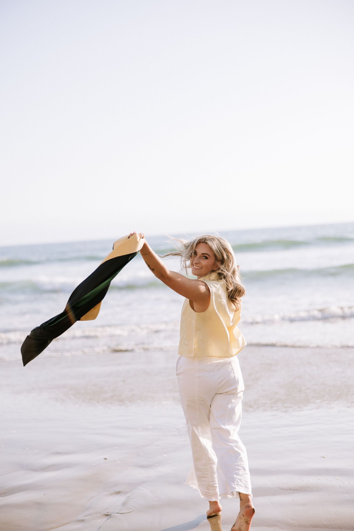 Young woman in yellow top and white pants waving graduation stole on sunny beach shoreline.