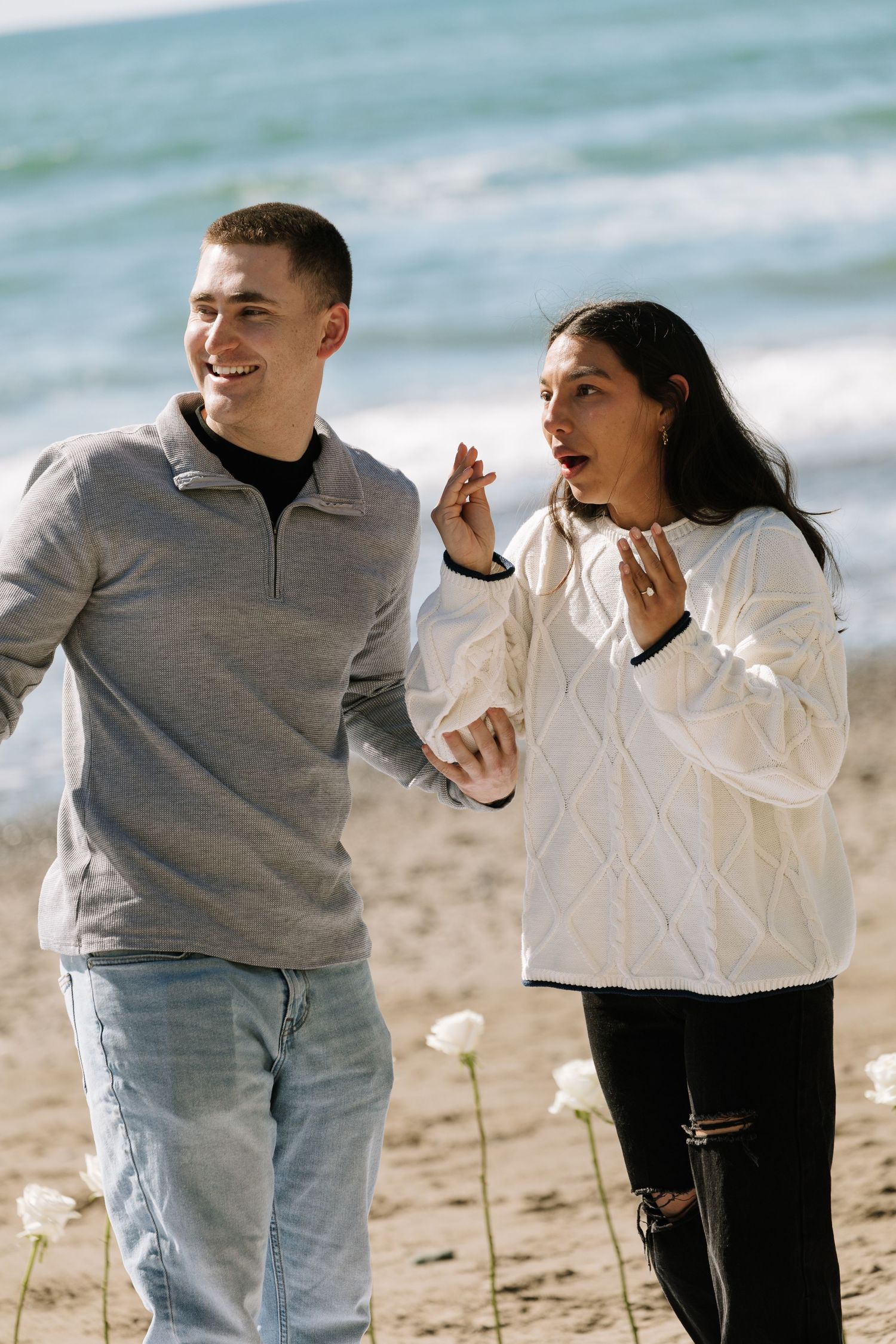 Excited newly engaged couple walking on the beach, woman admiring her engagement ring.