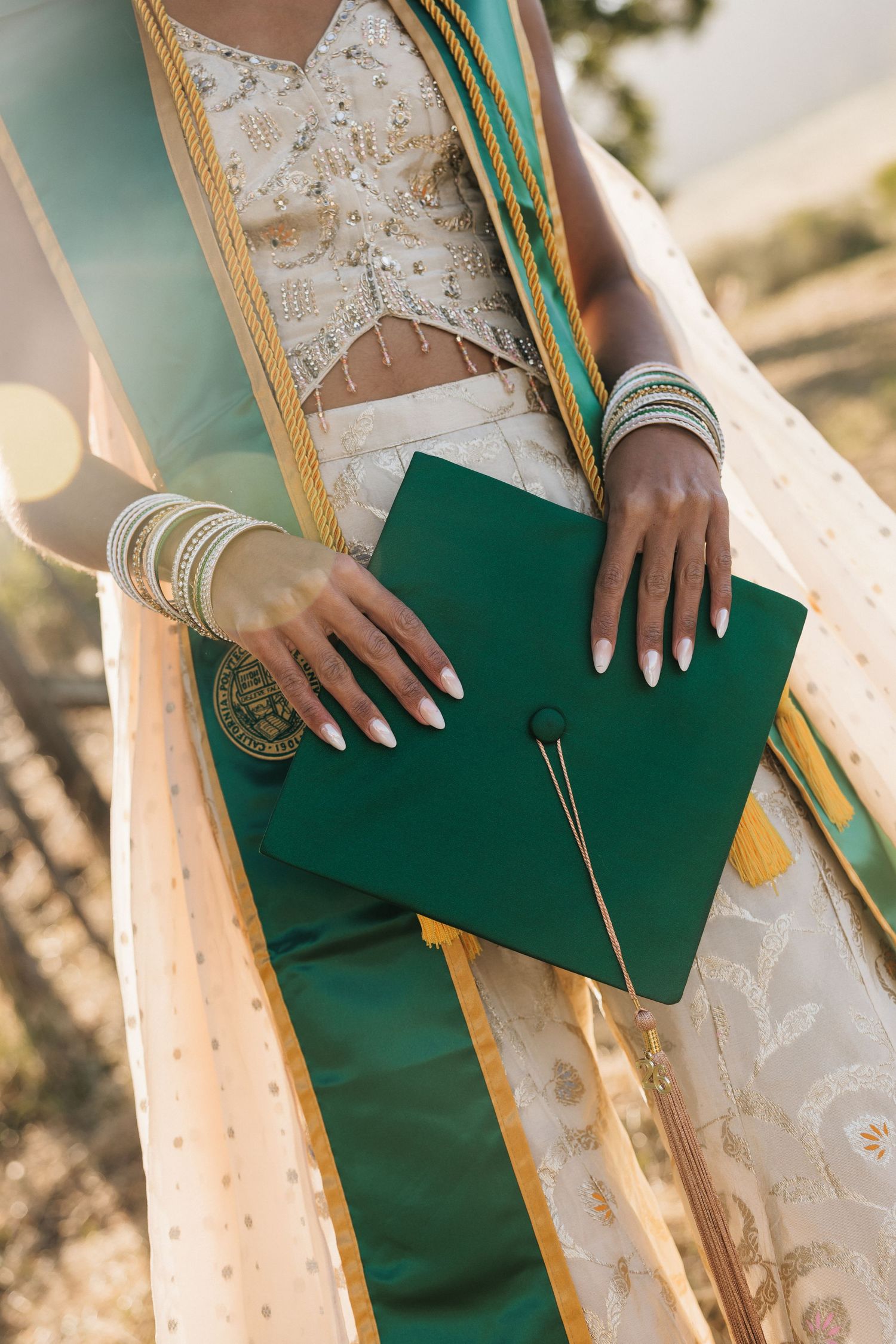 Close-up of hands holding green Cal Poly graduation cap with gold bangles and colorful graduation sash.