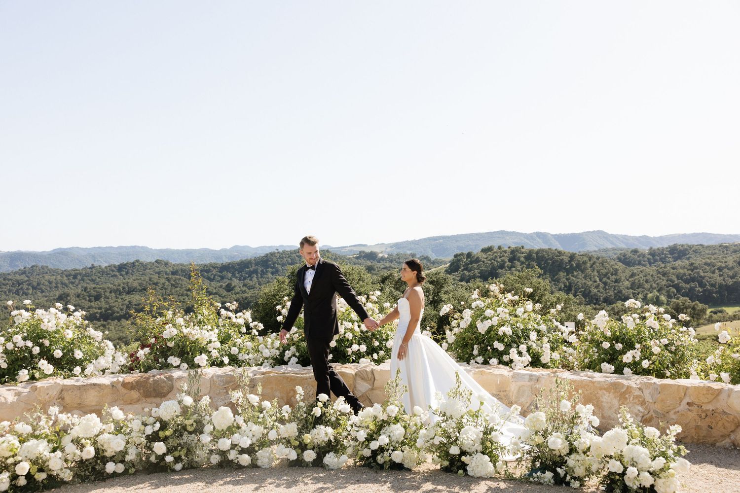 Bride and groom hold hands among white floral arrangements on a stone terrace overlooking rolling green hills.