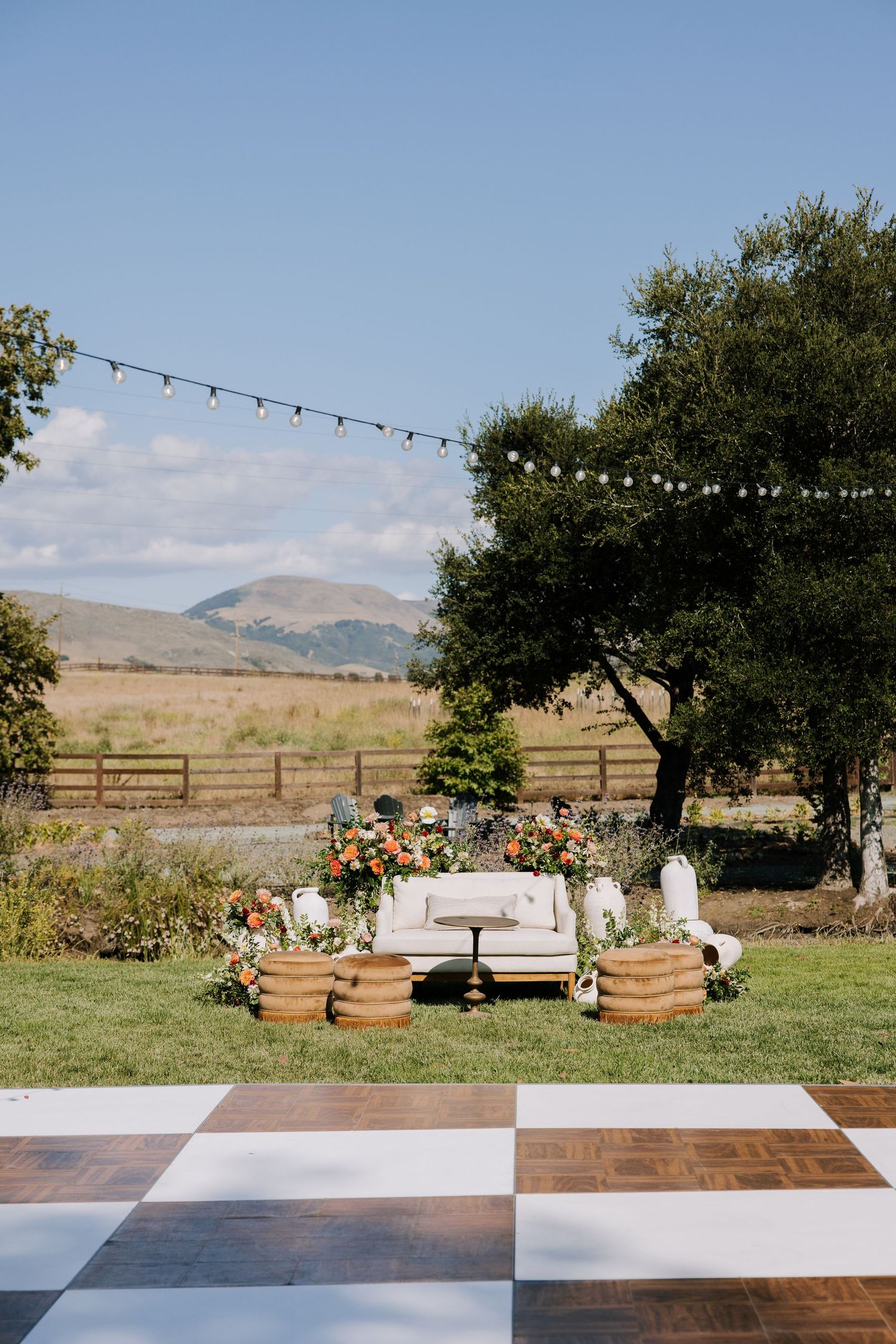 Elegant outdoor wedding lounge setup with checkered dance floor, floral arrangements, and string lights against mountain backdrop.