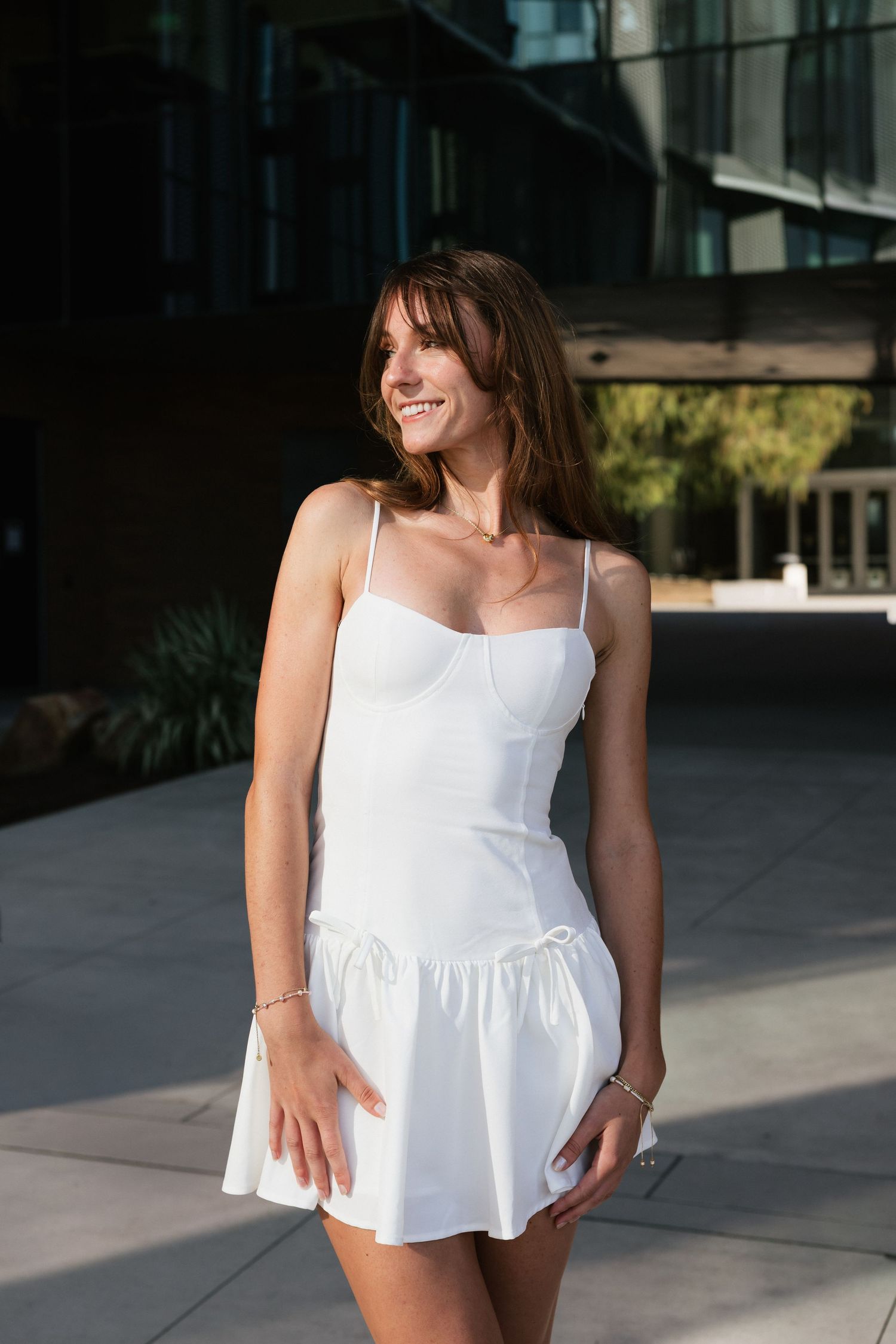 Young woman in white spaghetti strap mini dress smiling outdoors near modern glass building.