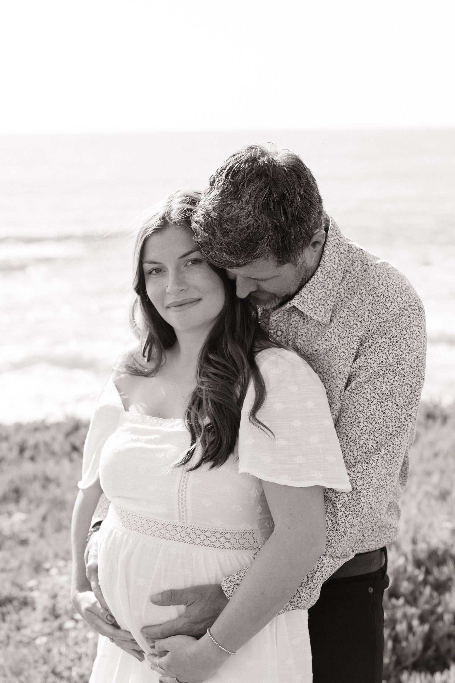 Black and white maternity portrait of couple embracing near ocean, man nuzzling pregnant womans head.