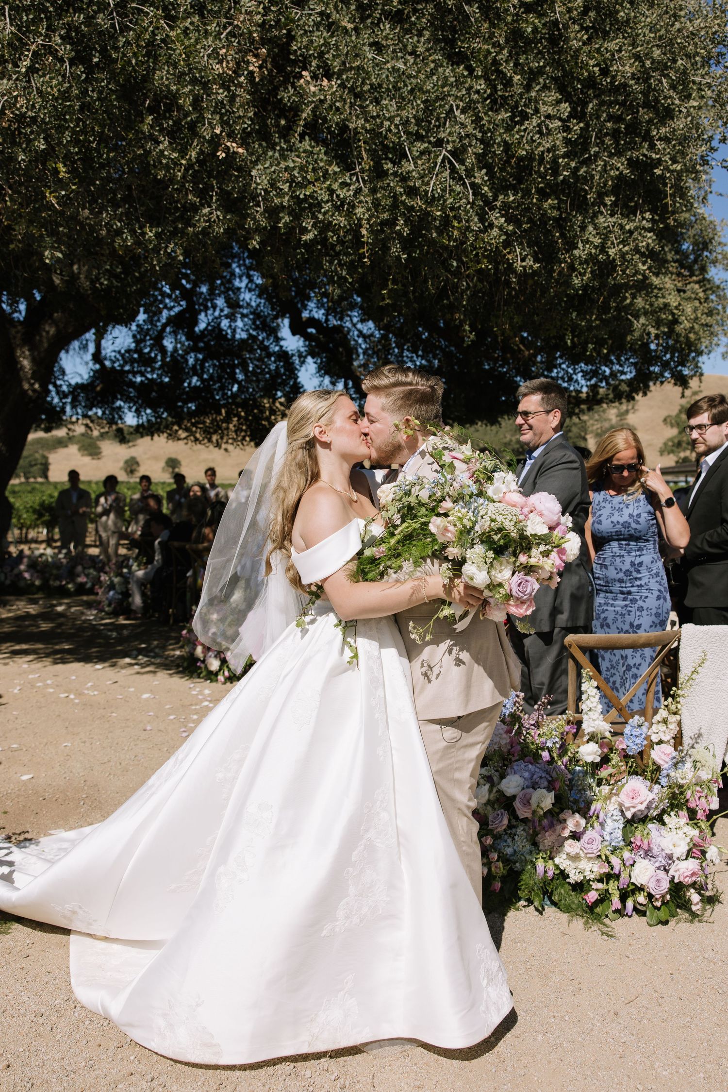Bride and groom share first kiss at outdoor wedding ceremony beneath oak tree with floral arch and bouquet.