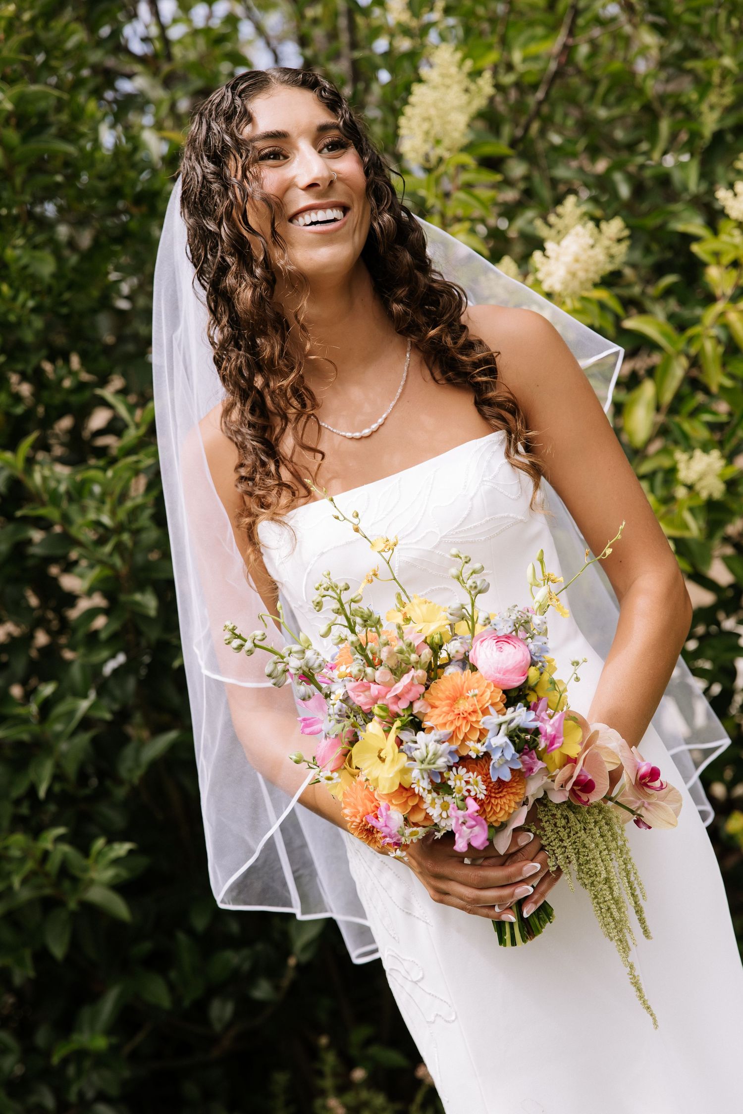 Smiling bride with curly hair holds a colorful bouquet while wearing a white strapless gown.
