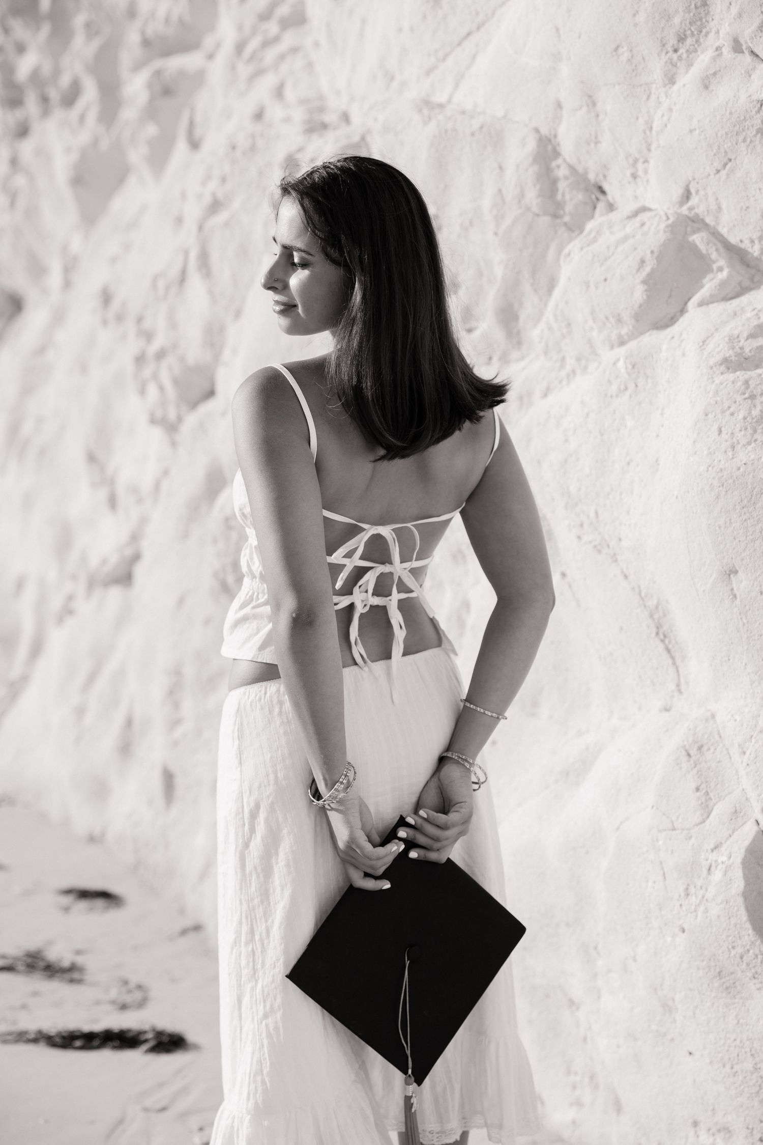Black and white portrait of woman in white dress holding graduation cap against white rock wall.