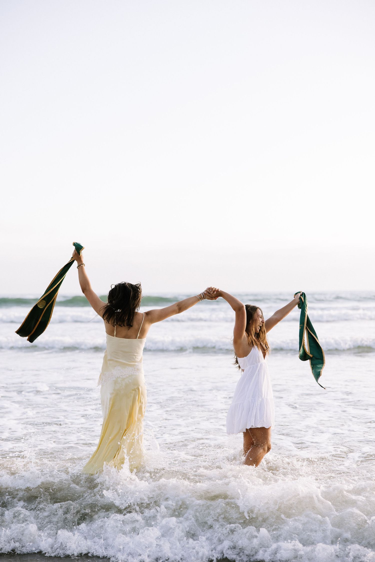 Two women in white and yellow dresses celebrating in ocean waves holding green stoles.