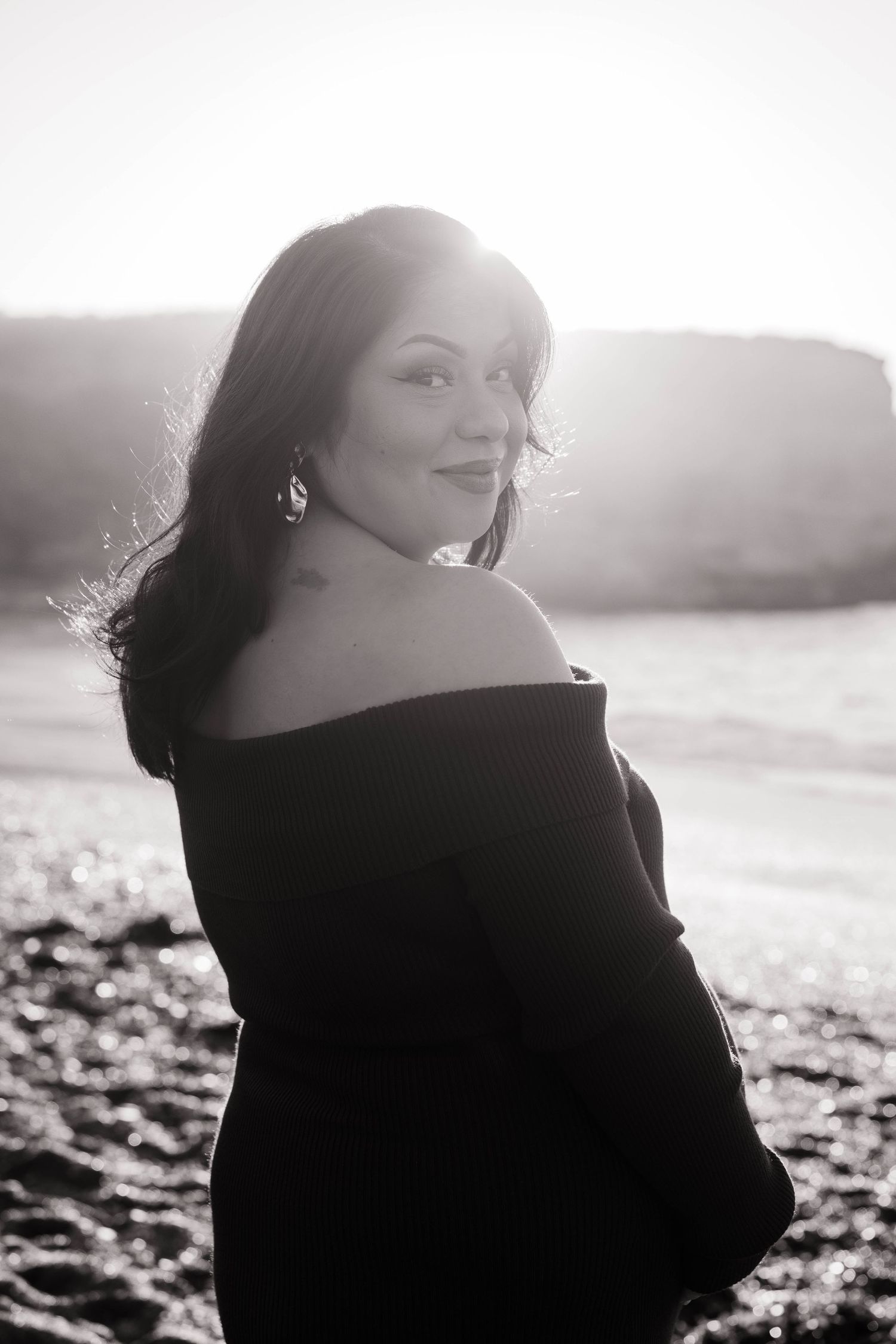 Black and white portrait of a pregnant woman in off-shoulder black dress smiling at beach during golden hour.