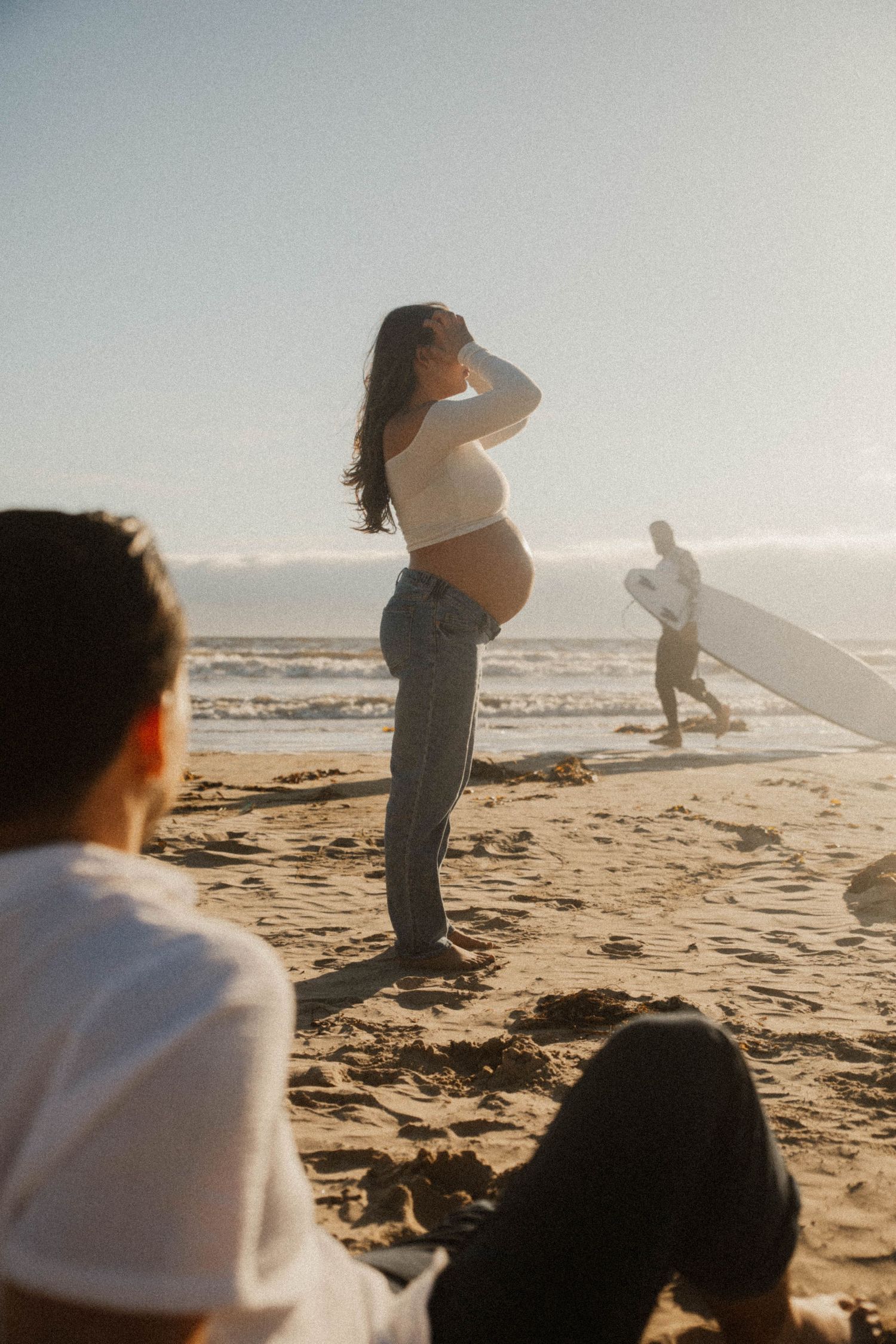Pregnant woman in white crop top and jeans standing on sunny beach while surfer walks in background.
