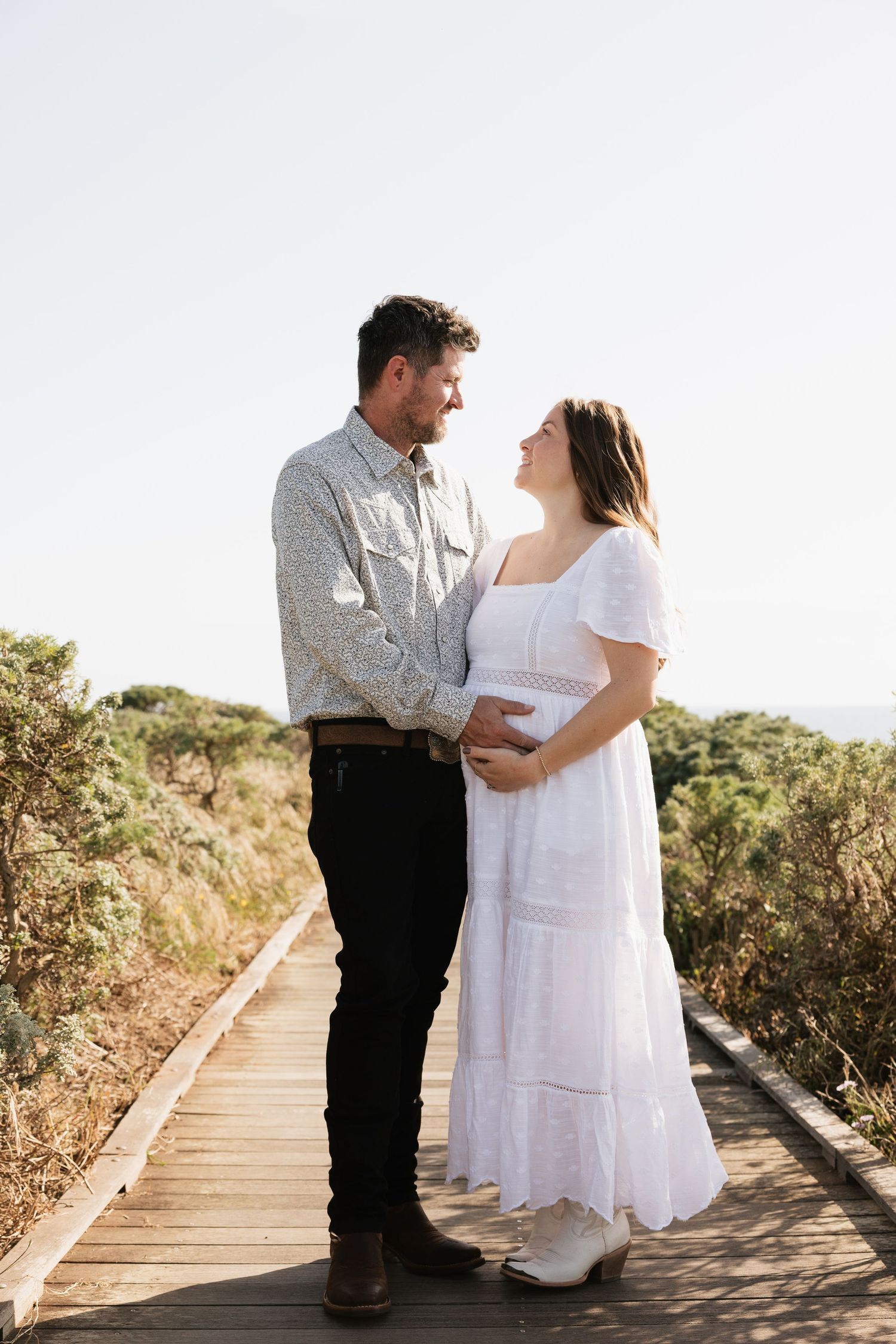 Expecting couple standing on a wooden boardwalk, hands on baby bump, smiling warmly.
