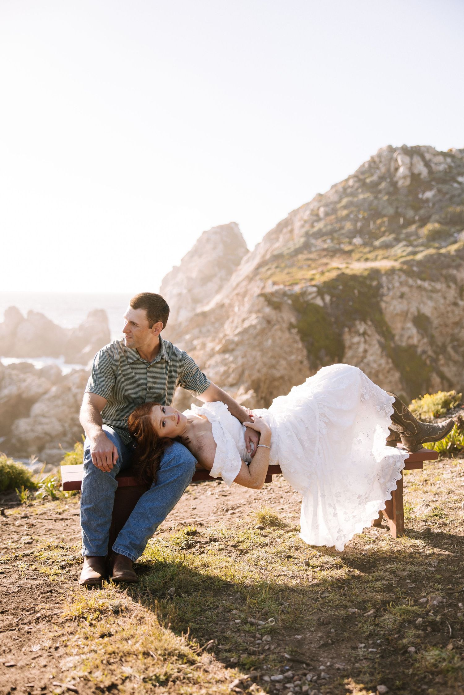 Couple relaxing on a bench at golden hour, woman lying across mans lap with ocean cliffs behind.
