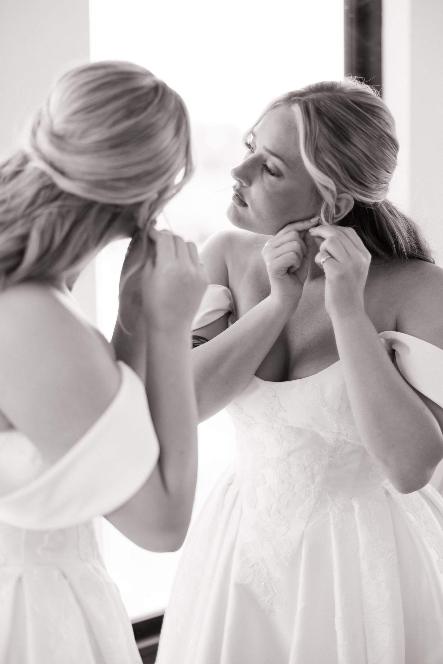 Black and white photo of bride adjusting earring while looking in mirror in white wedding gown.